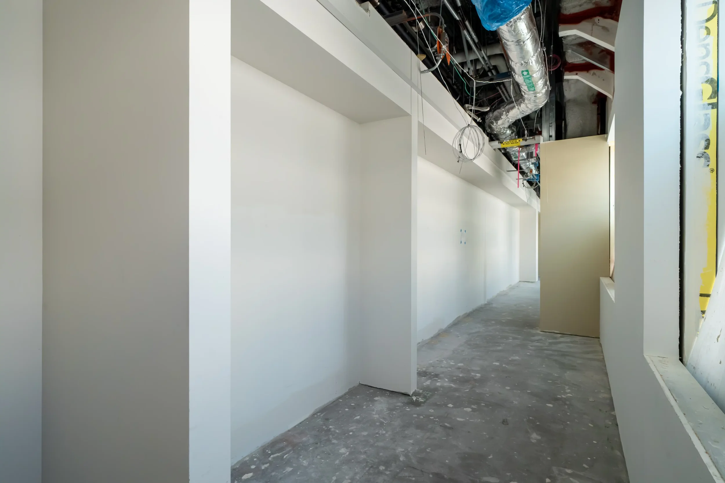Interior view of a building under construction with white painted walls, exposed ceiling with ducts and wiring, and a doorway on the right.