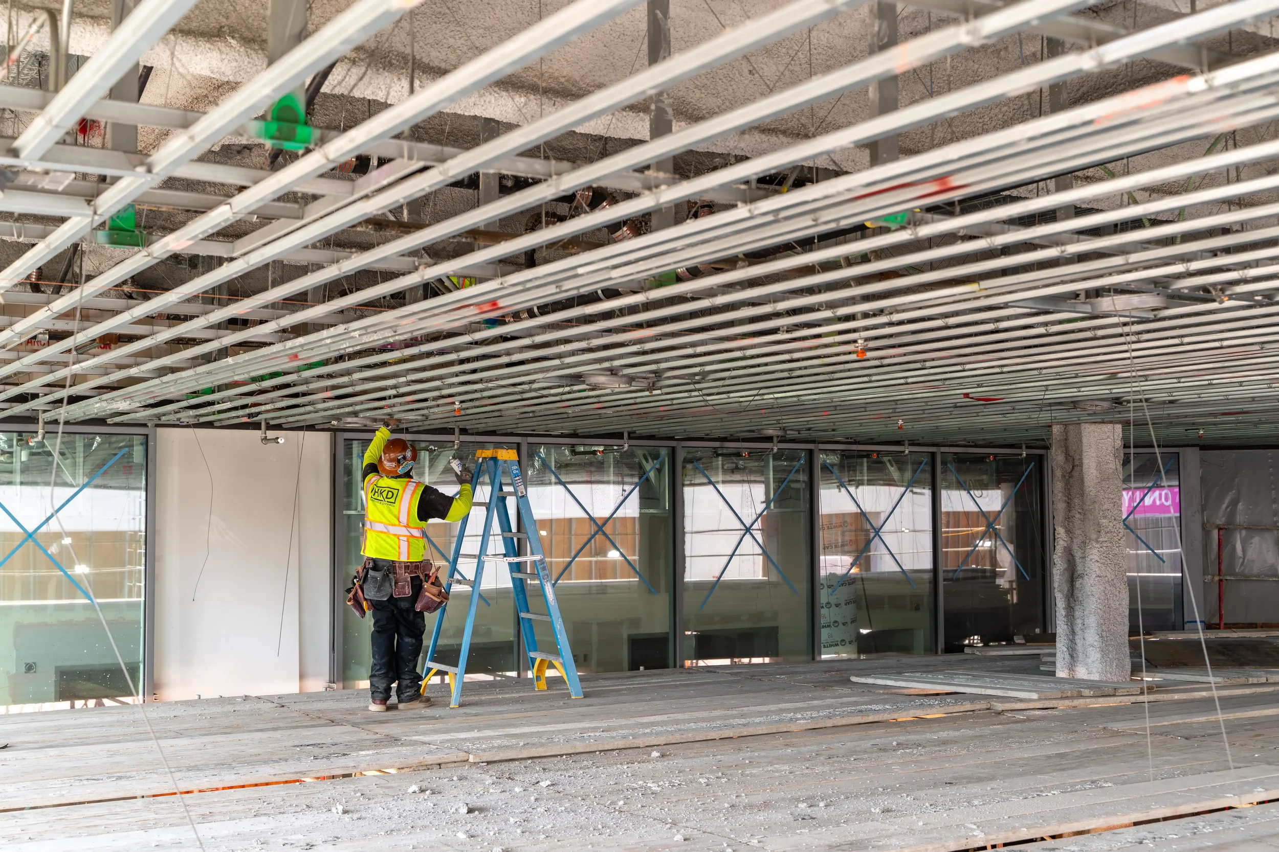 Construction worker with safety helmet and high visibility vest working on ceiling framework inside a building under construction.