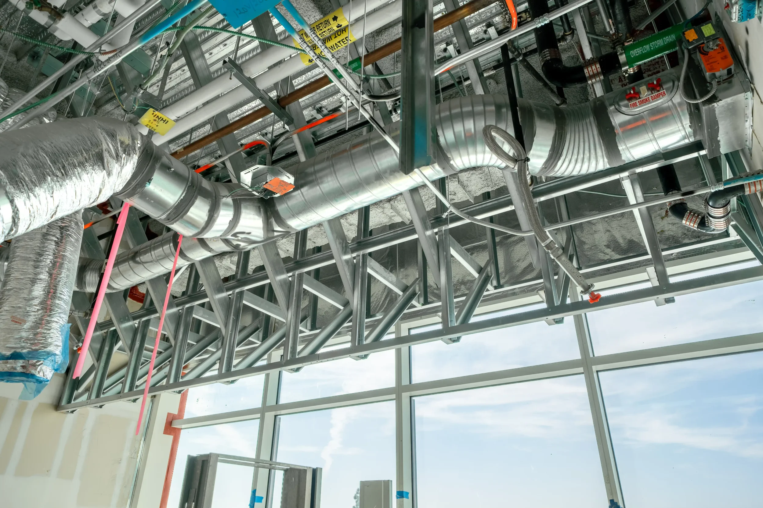 Construction site interior with exposed metal framework, ventilation ducts, and pipes, some with warning labels, against a large window showing a cloudy sky outside.
