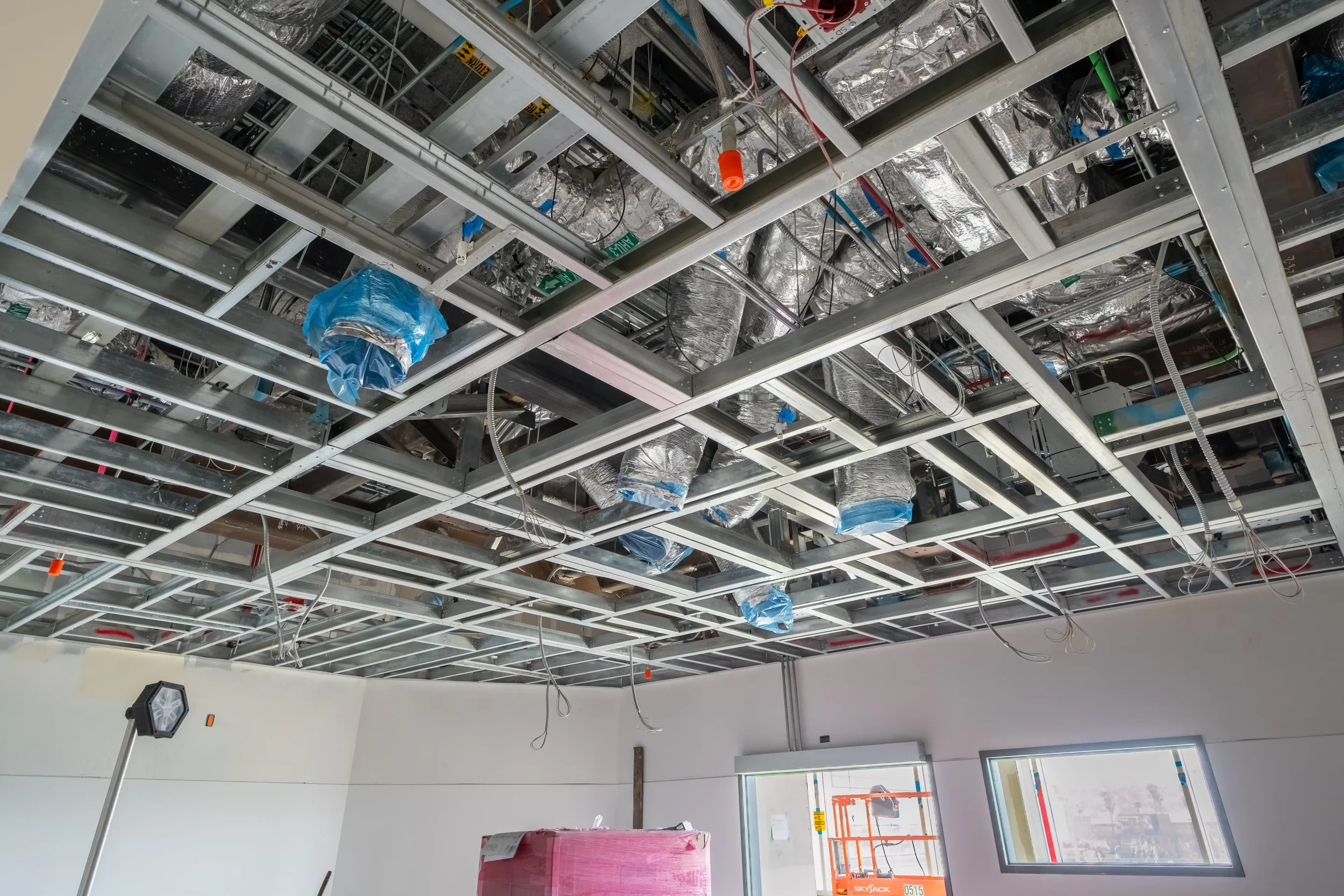Construction site ceiling with metal framing and silver insulated ducts, some covered with blue plastic, and electrical wires hanging down. There is a white wall with windows and an exit sign, and construction tools on the floor.