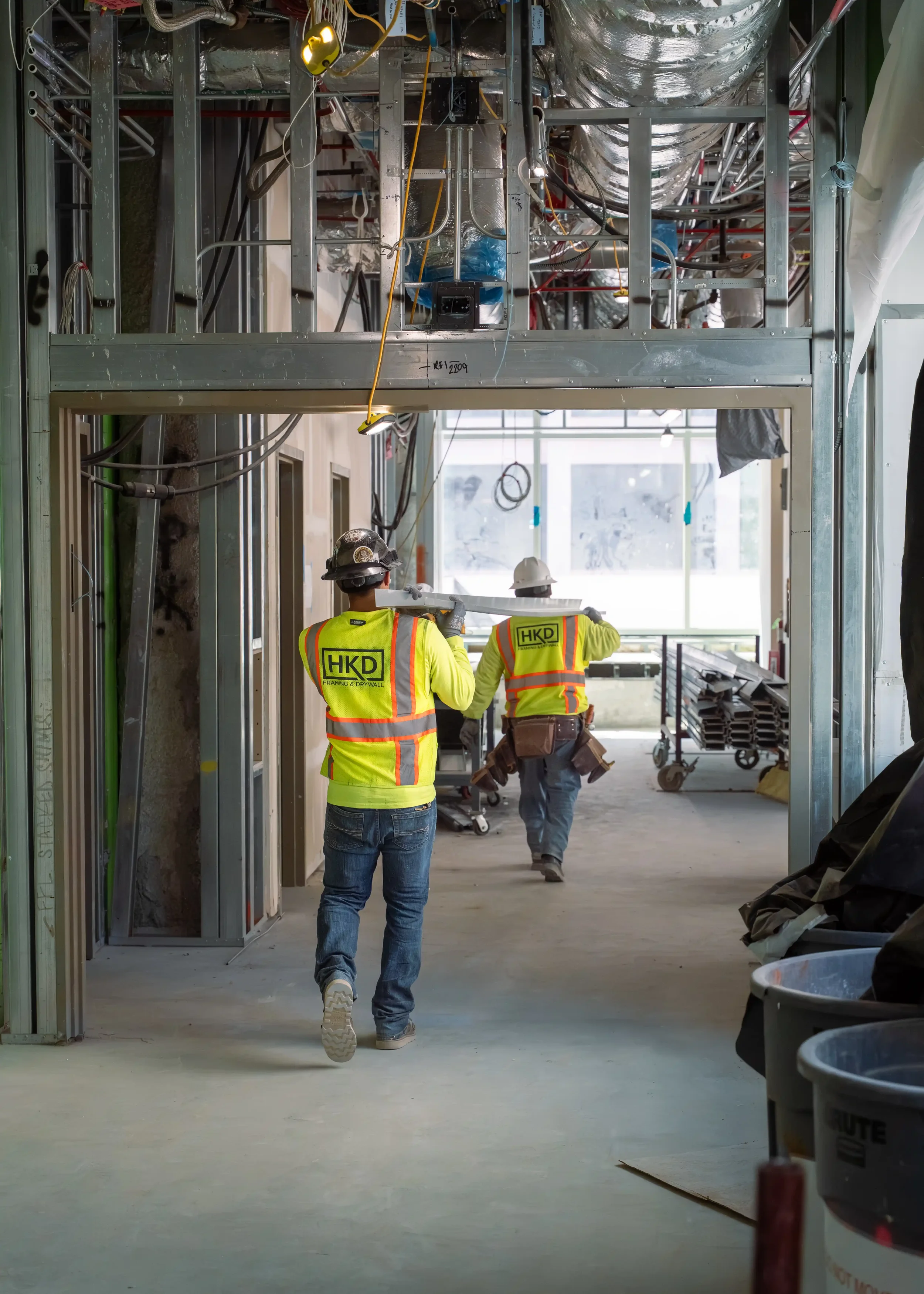 Construction workers in yellow safety vests and helmets walking through an unfinished building with exposed metal framing, electrical wiring, and construction materials.