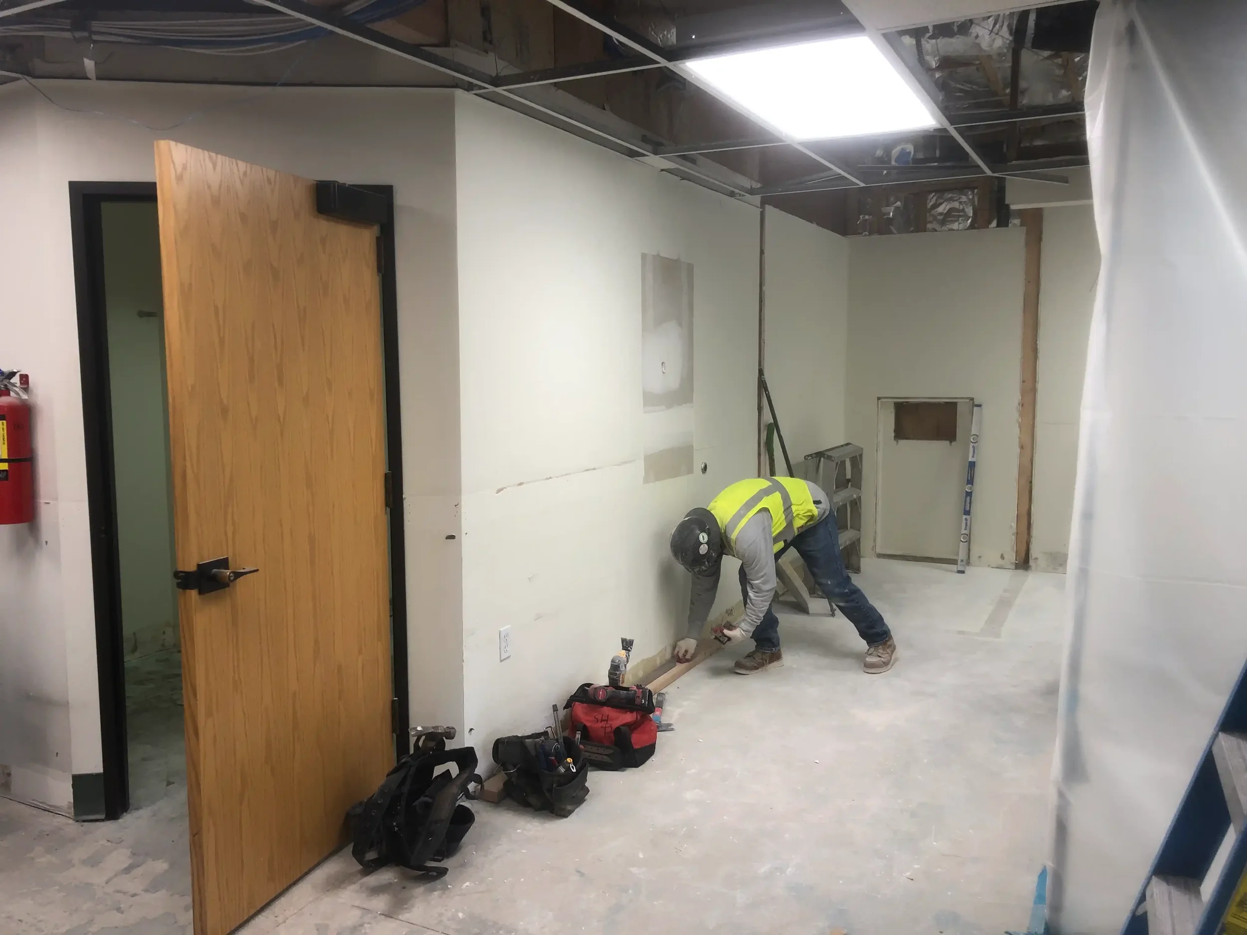 Construction worker in safety gear working on wall in unfinished interior space with tools and equipment on the floor.
