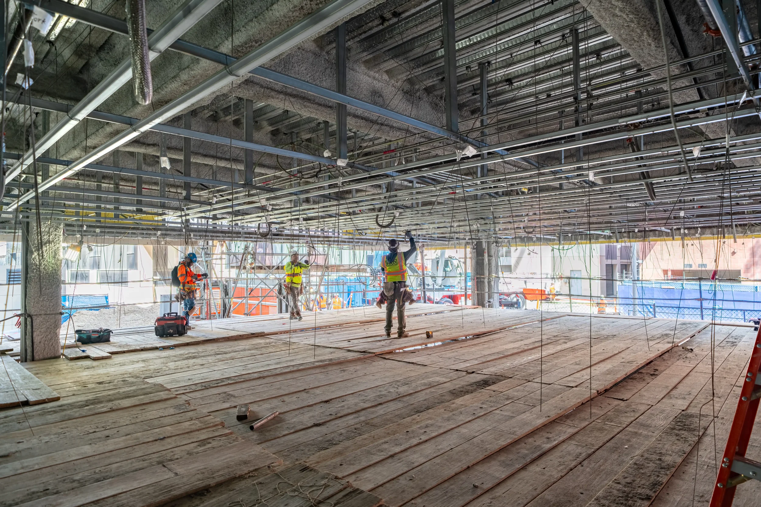 Construction workers in safety vests and helmets working on a building site with exposed ceiling framework and wooden flooring.