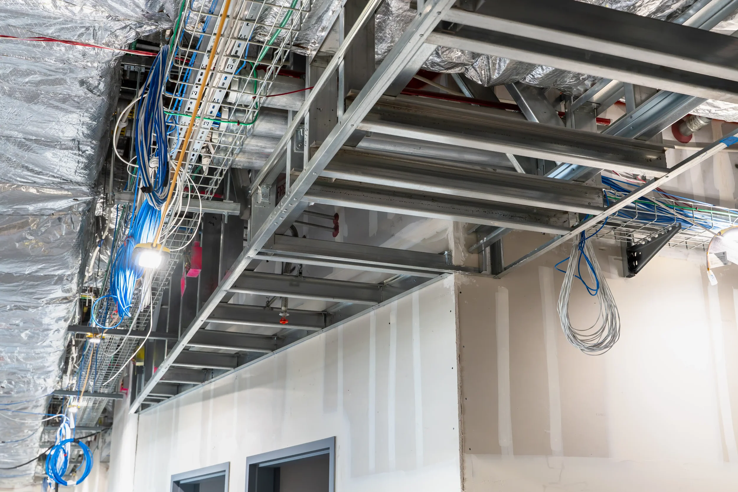 Inside a building under construction, metal framing and electrical wiring are installed along the ceiling.