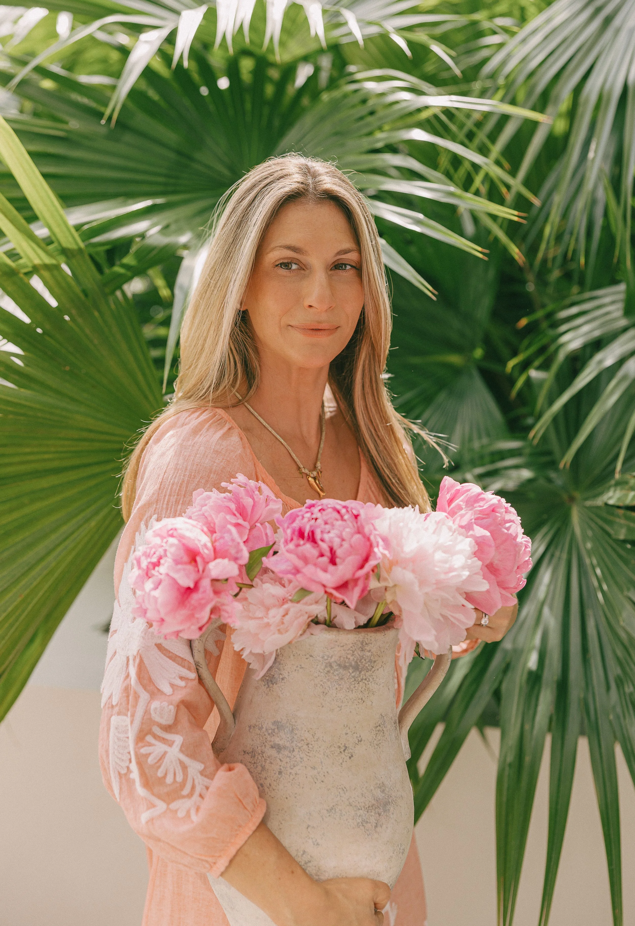 A woman with long blonde hair holding a white vase with pink peonies, standing in front of lush green palm plants.