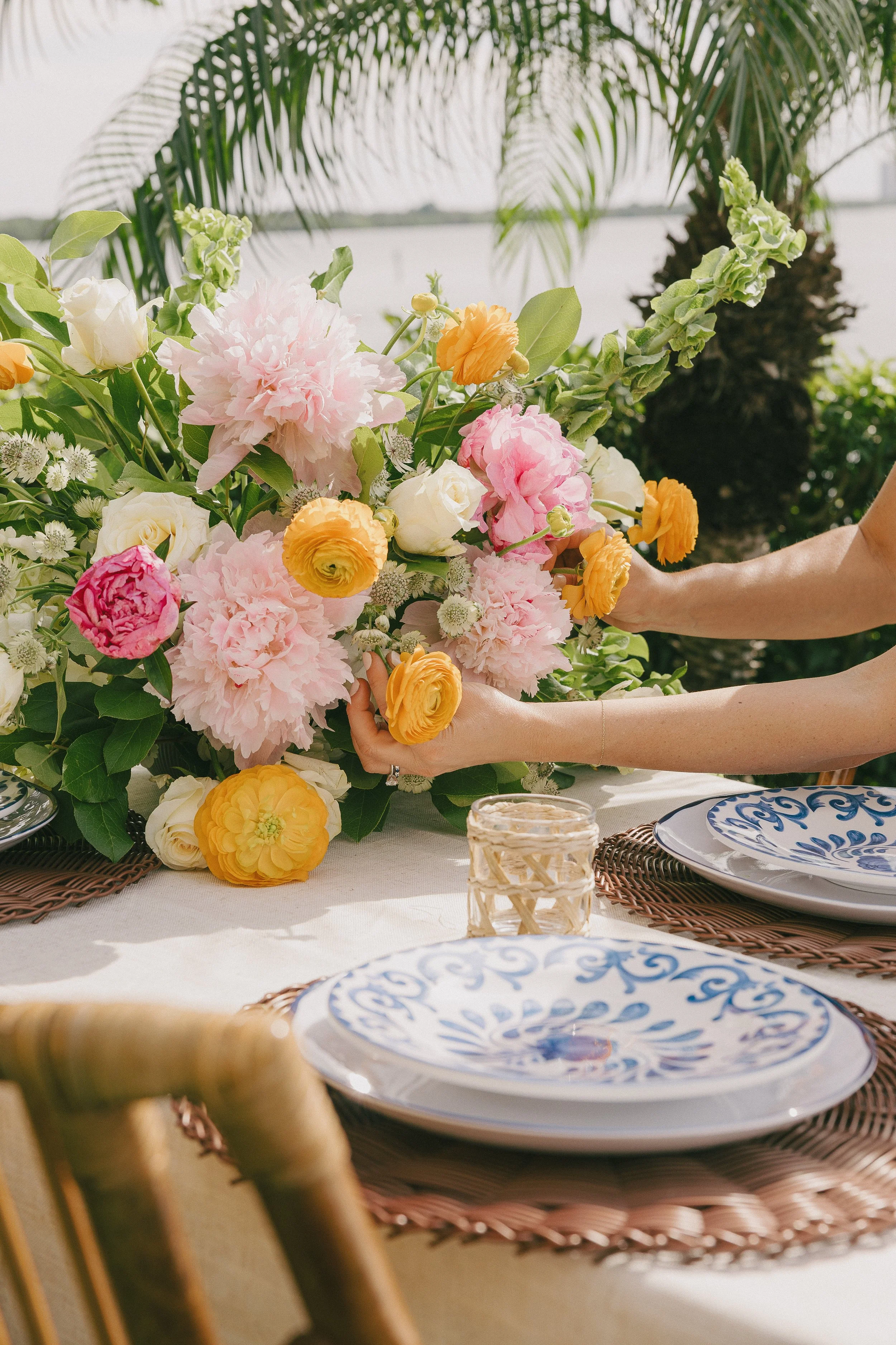 A table setting outdoors with a large floral arrangement of pink, yellow, white, and orange flowers, with plates and glasses on a woven placemat, and palm trees in the background.