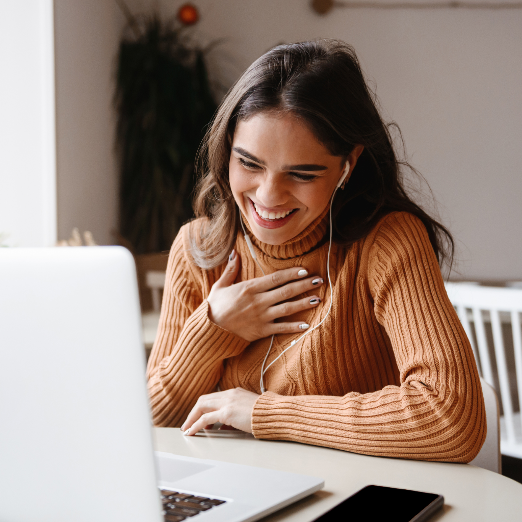 A woman with long brown hair smiling and wearing a brown turtleneck sweater while engaged in a video call or online activity, with a laptop on the table in front of her.