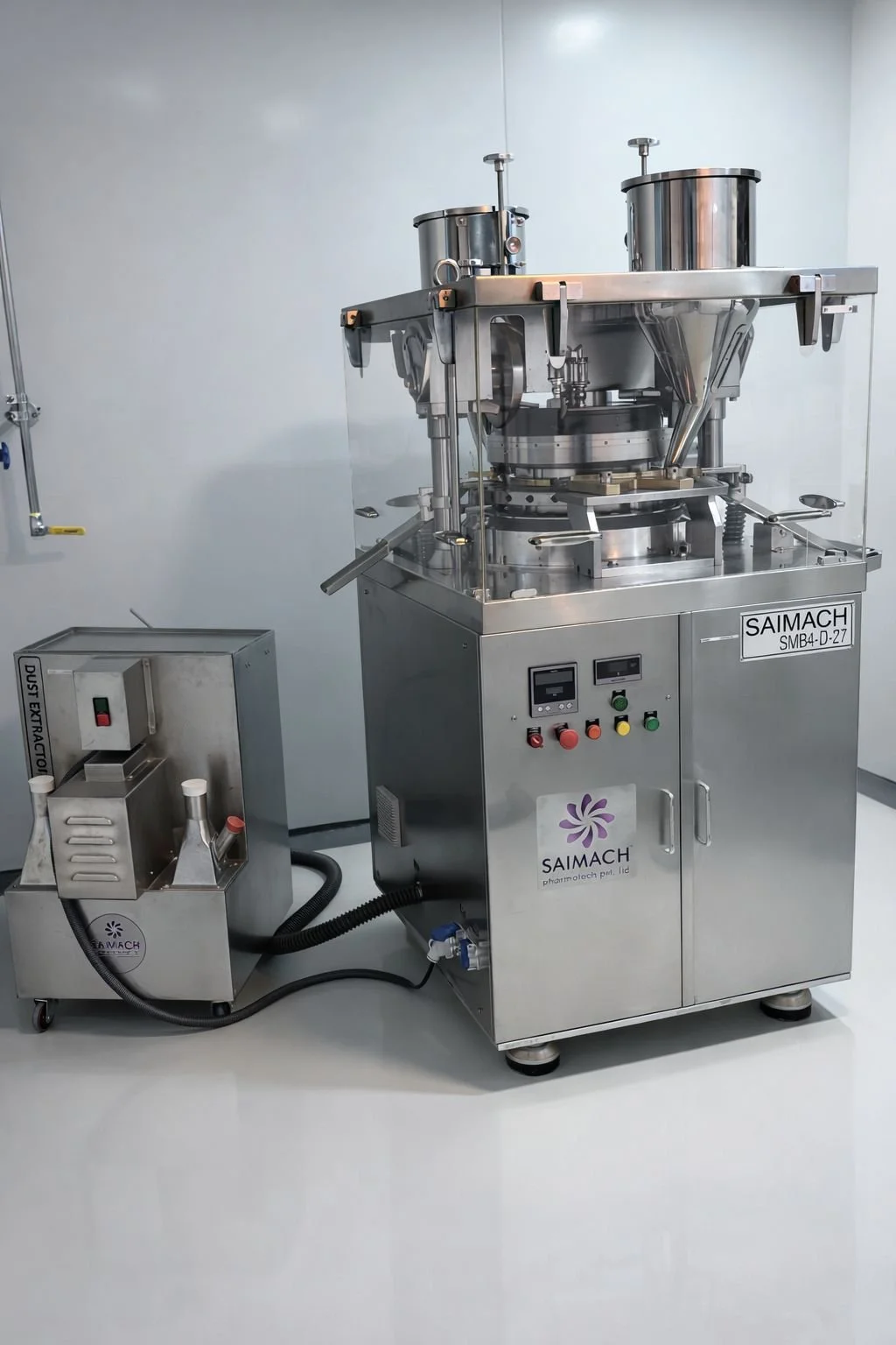A stainless steel industrial machine with controls, labeled 'SAIMACH,' used for pharmaceutical or manufacturing processes. It is connected to a smaller attached device via a black hose, with a clean, minimal laboratory or manufacturing room backgroun