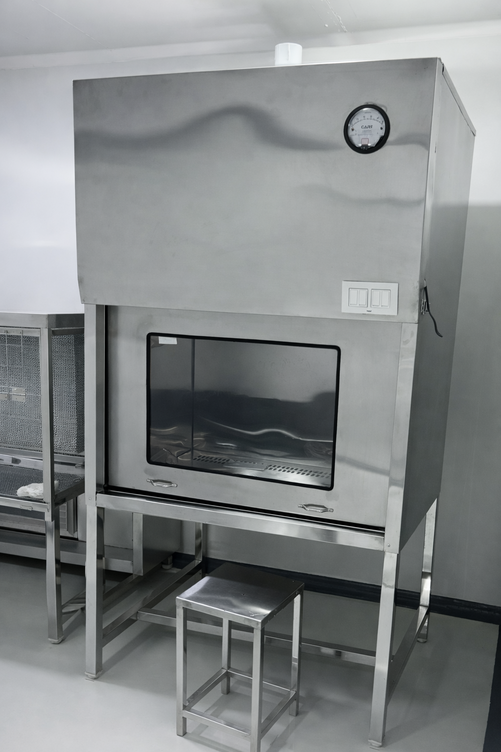 A stainless steel laboratory fume hood with a glass window, a gauge on top, and a small stool in front.