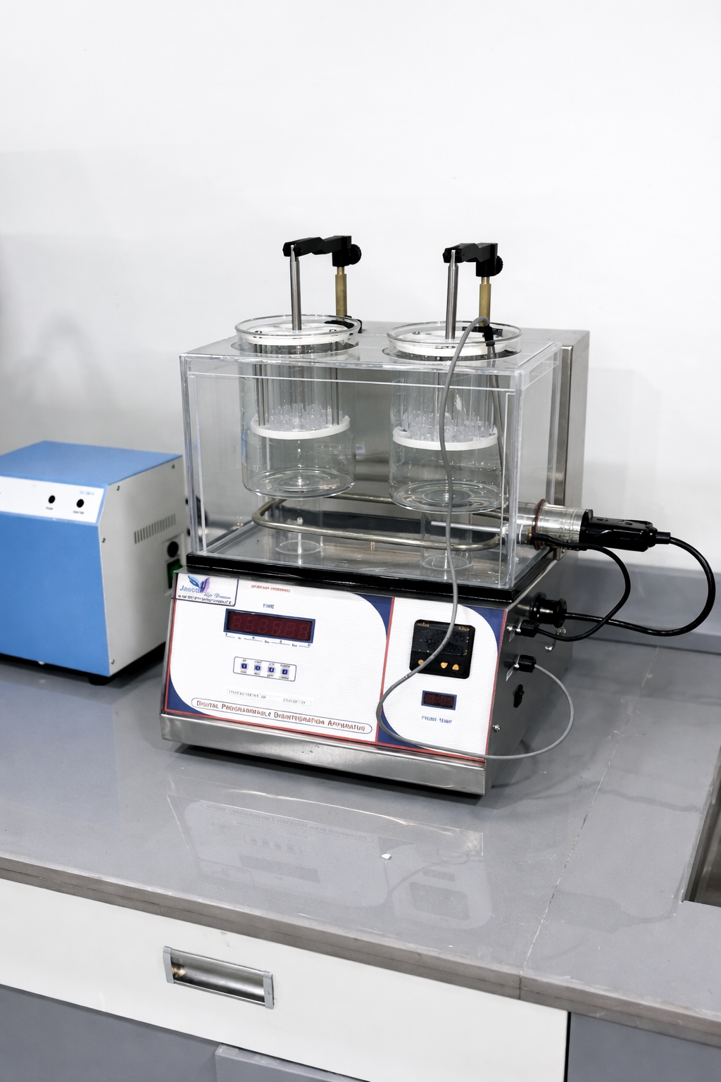 Laboratory equipment with two glass containers inside a transparent protective shield, connected to a machine with control buttons and a digital display, used for scientific experiments or testing, placed on a gray work surface.
