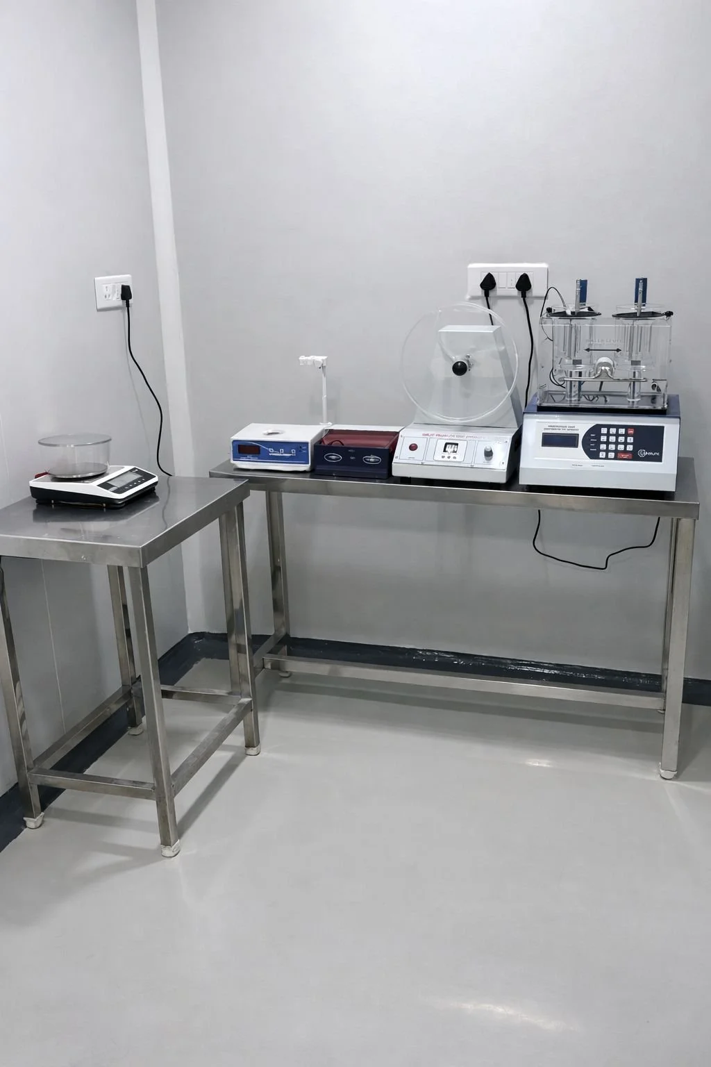 Laboratory workspace with steel tables, scientific equipment including centrifuge, scales, and other devices, against a white wall.