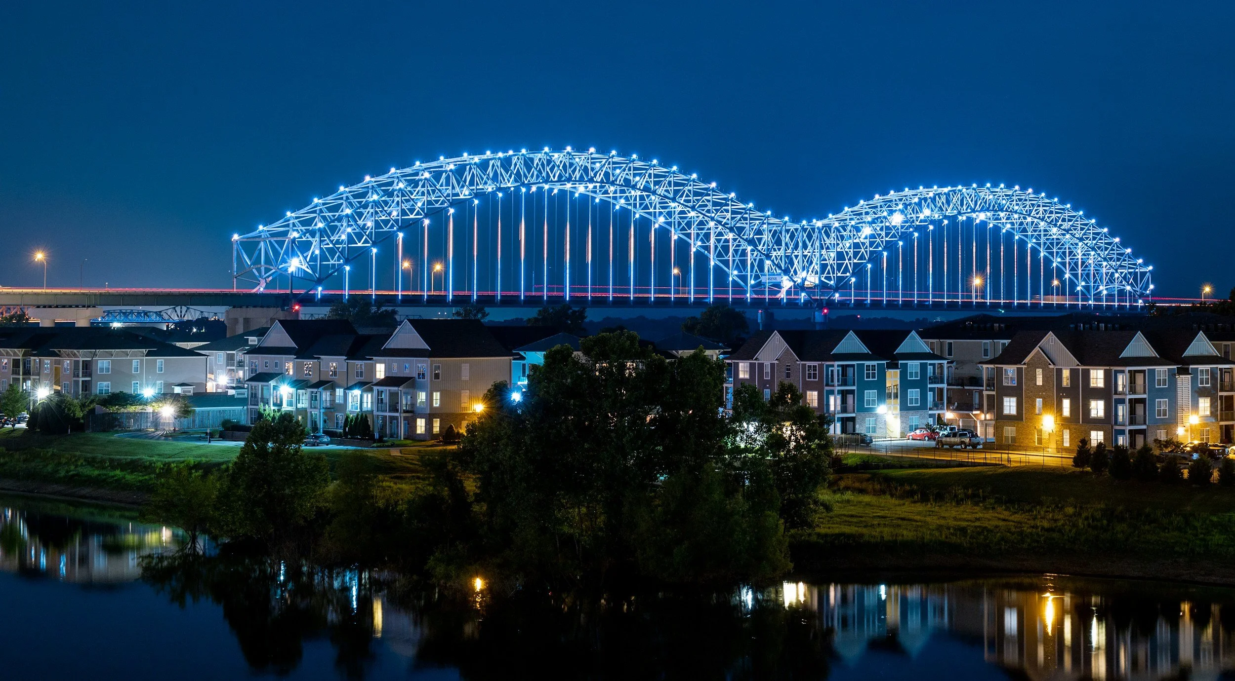 A brightly lit blue arch bridge at night over a body of water with a residential area with lit windows and trees in the foreground.