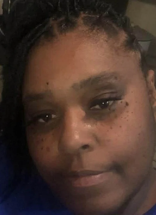 Close-up of a person's face with dark skin, short curly hair, and visible freckles, looking at the camera with a neutral expression.
