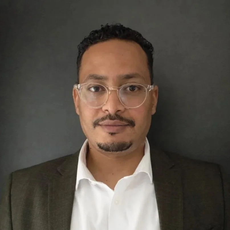 A man with short curly hair, glasses, and a goatee, wearing a dark blazer and white shirt, standing against a dark background.