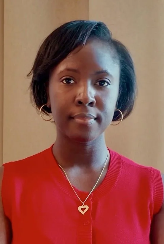 A woman with short black hair wearing a red sleeveless top, hoop earrings, and a heart-shaped pendant necklace, standing indoors against a beige background.