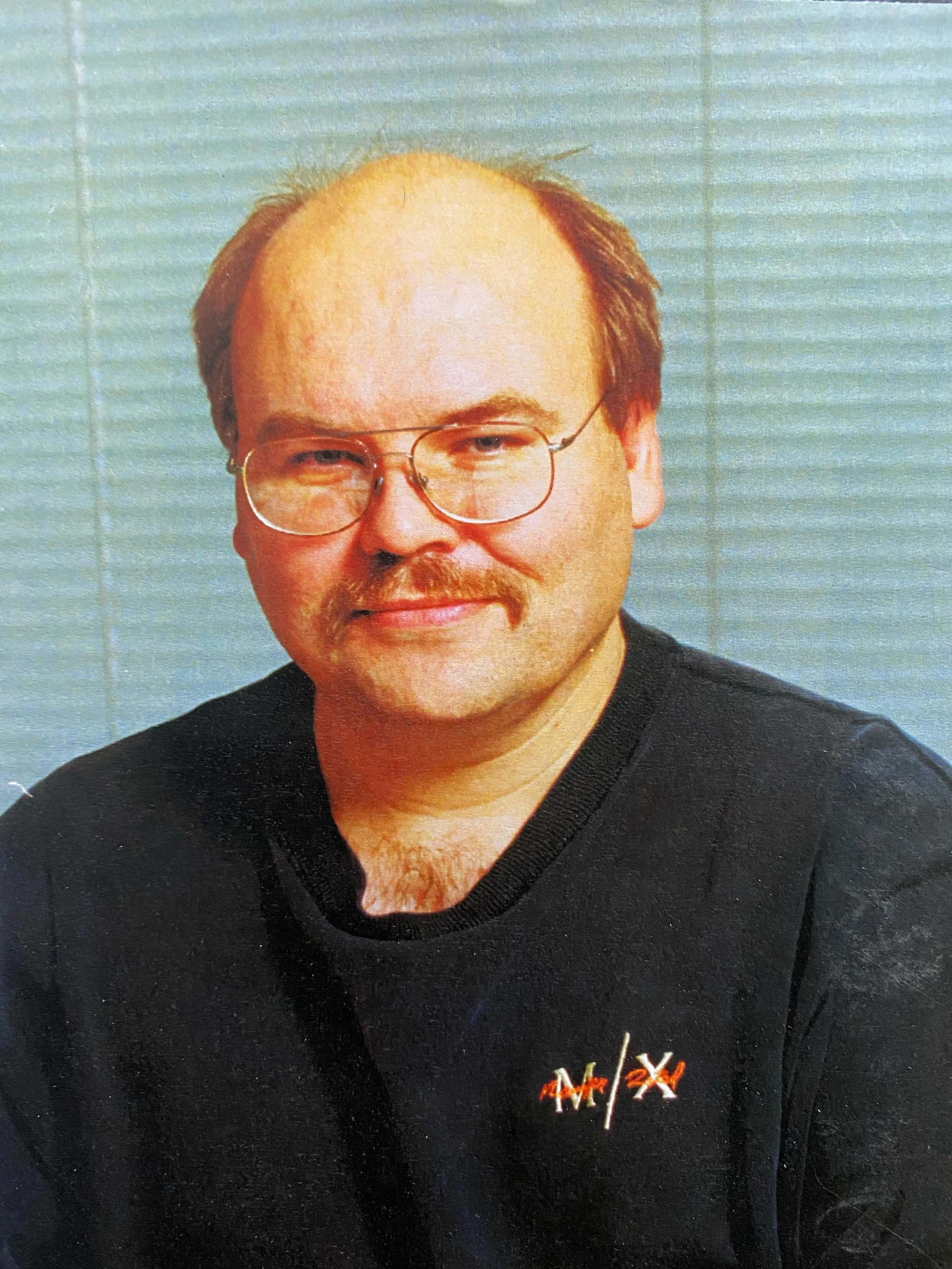 A man with glasses, short hair, and a mustache wearing a black shirt with a logo that reads 'MX'.