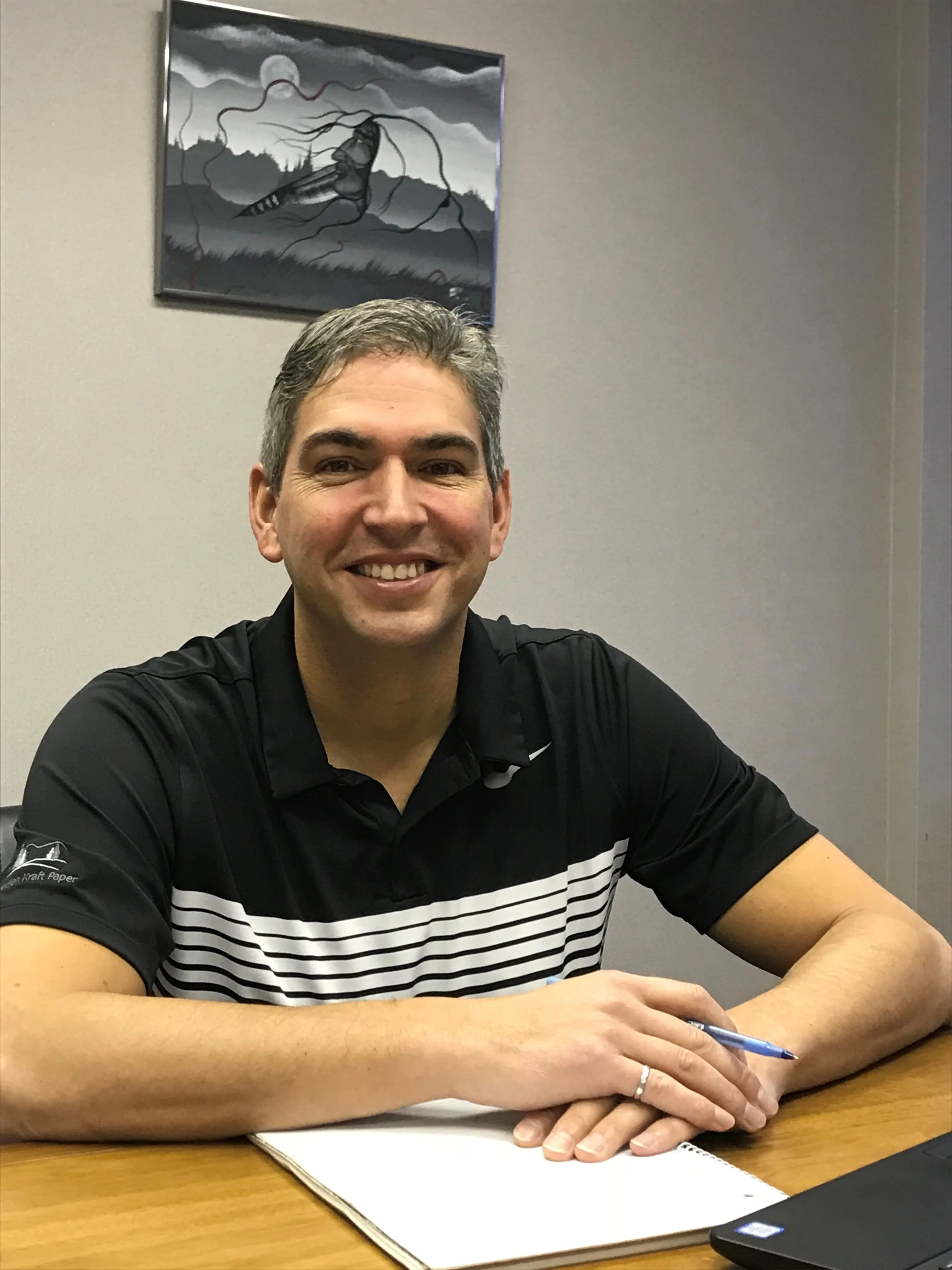 A smiling man with short gray hair sitting at a wooden table in front of a notepad and pen, wearing a black polo shirt with white horizontal stripes, with a painting of a landscape featuring mountains, trees, and a large insect hanging on the wall behind him.