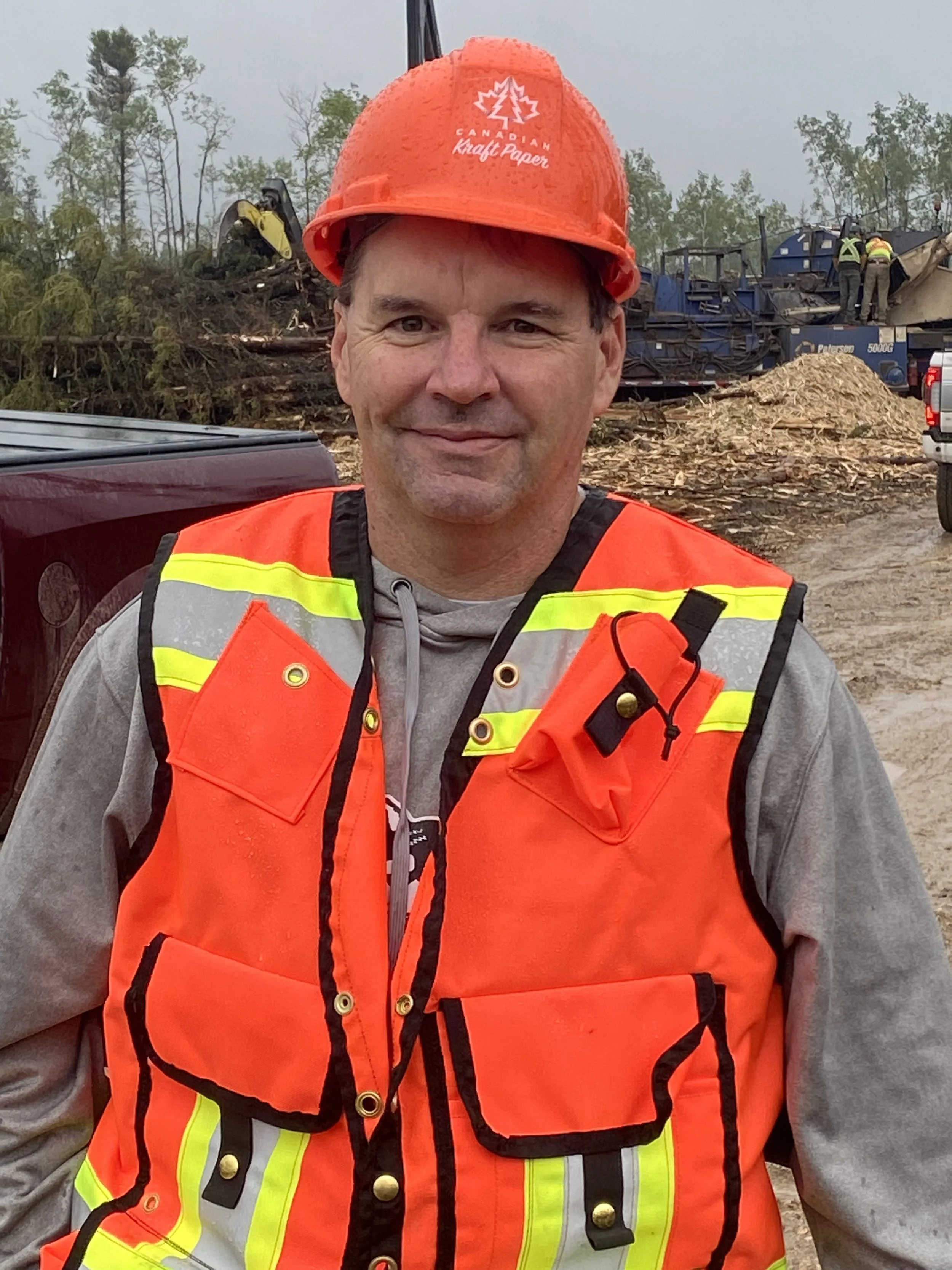 A man wearing a bright orange safety vest and a red construction helmet standing outdoors at a construction site with heavy machinery and debris in the background.