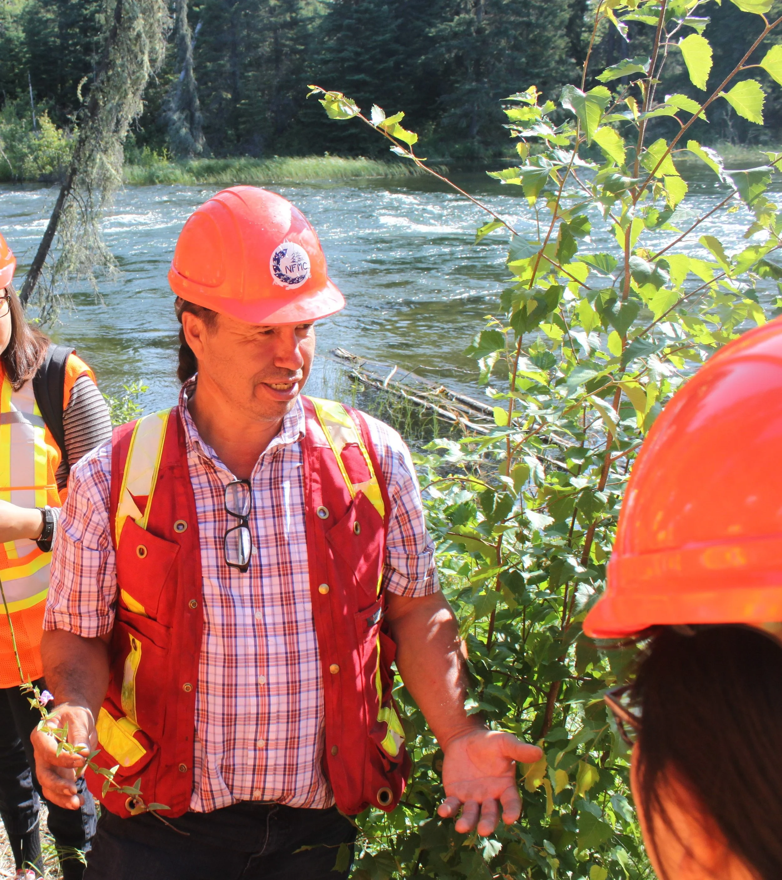 A man wearing a red safety vest and orange hard hat talking to a group of people near a riverbank, with trees and water in the background.