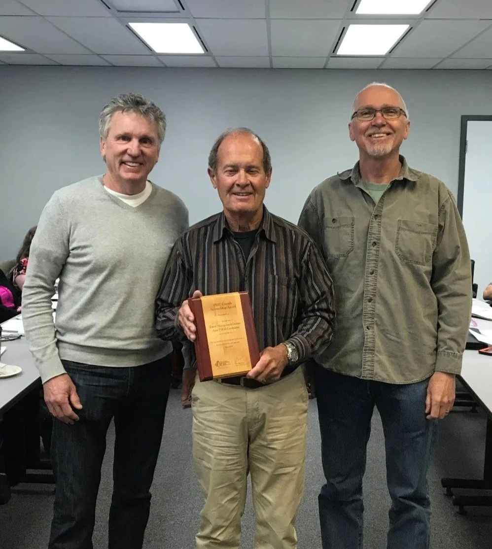 Three men standing together in a conference room, with one man in the center holding a plaque or award. All three are smiling.