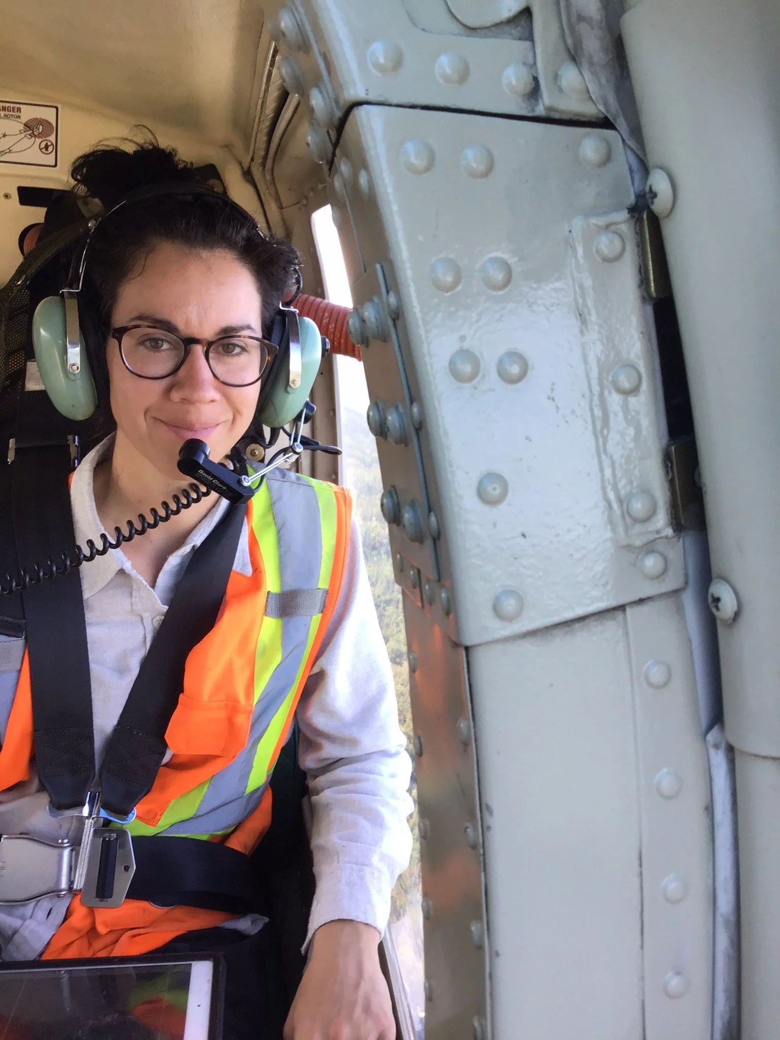 A woman flying in a helicopter wearing headphones, a safety vest, and glasses.