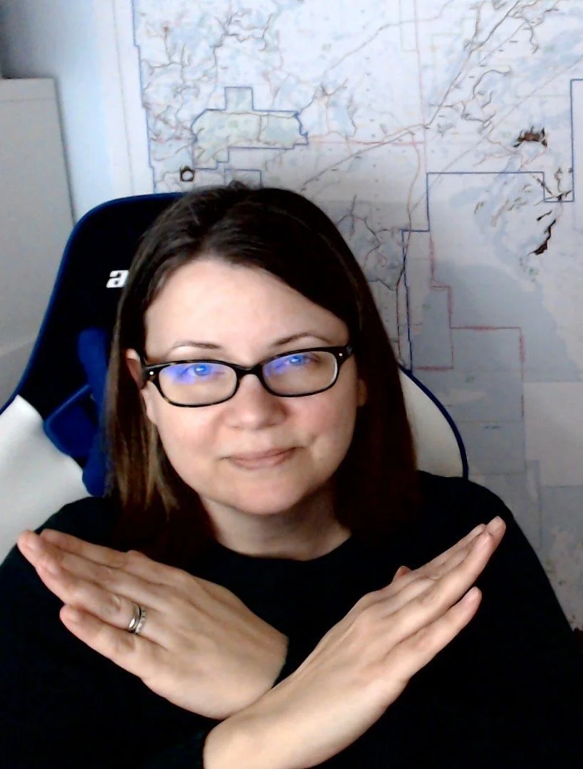 A woman with glasses poses with her arms crossed in front of her chest, in front of a wall with a map. She is wearing a black top and has shoulder-length brown hair.
