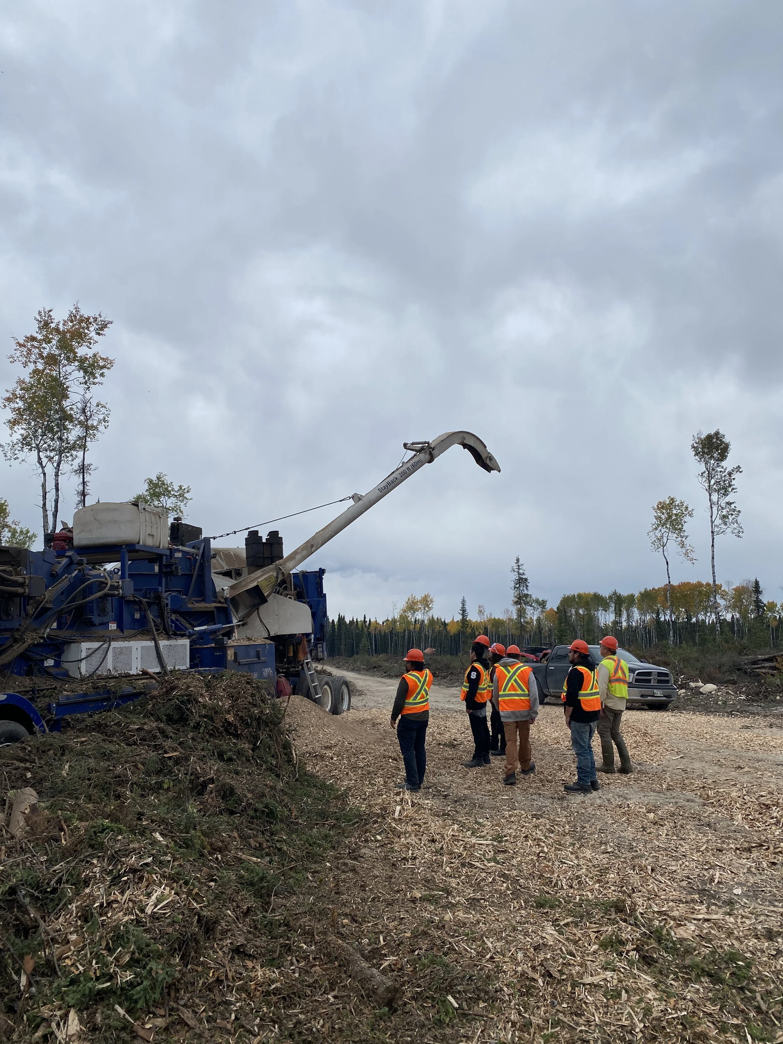 Six construction workers wearing safety vests and helmets stand on a dirt road watching a large wood chipper in operation, processing branches into wood chips, with a forest in the background and a cloudy sky overhead.