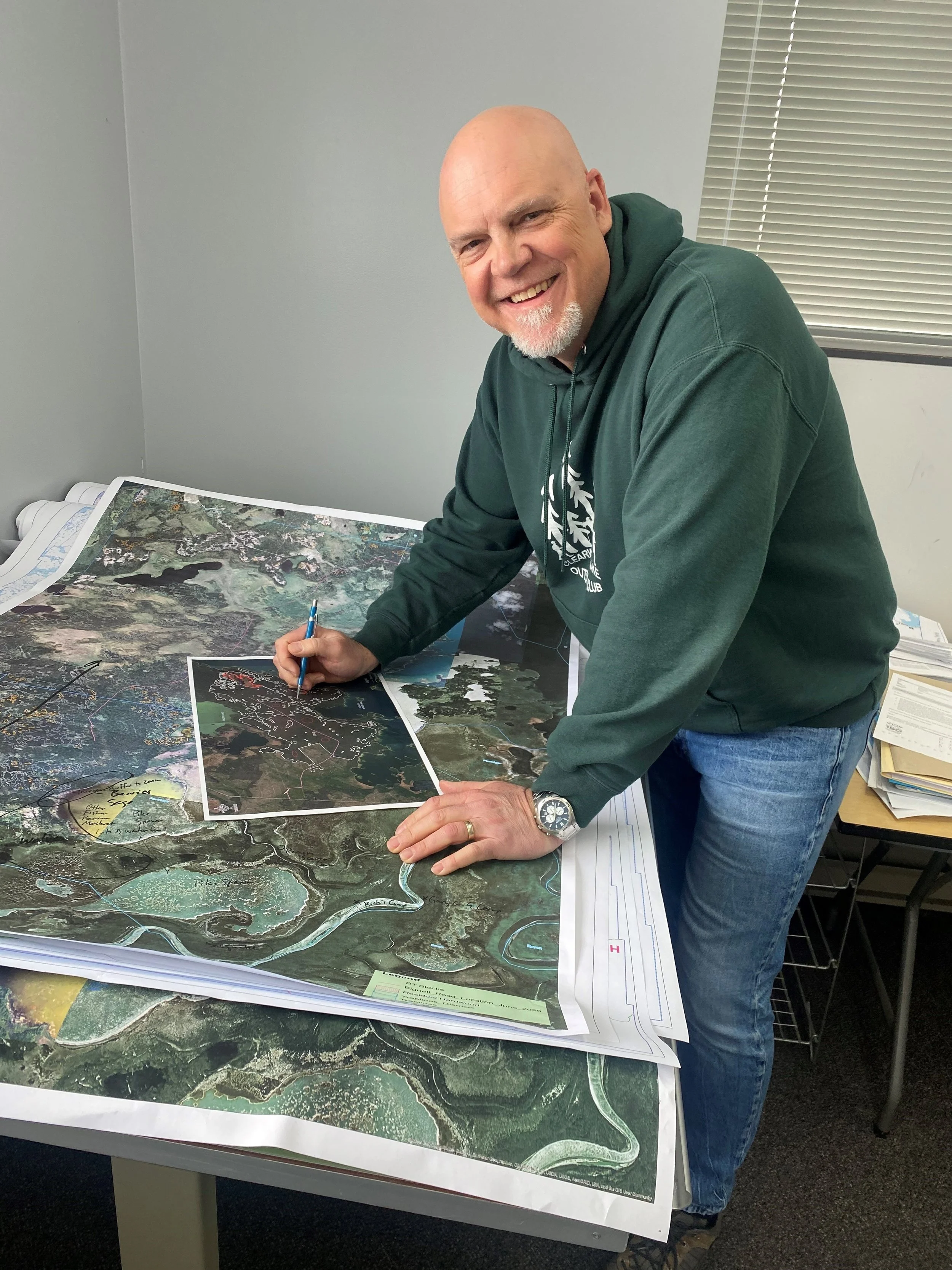 A man in a green hoodie and jeans is smiling while signing an aerial map of a landscape on a table filled with large maps and documents.