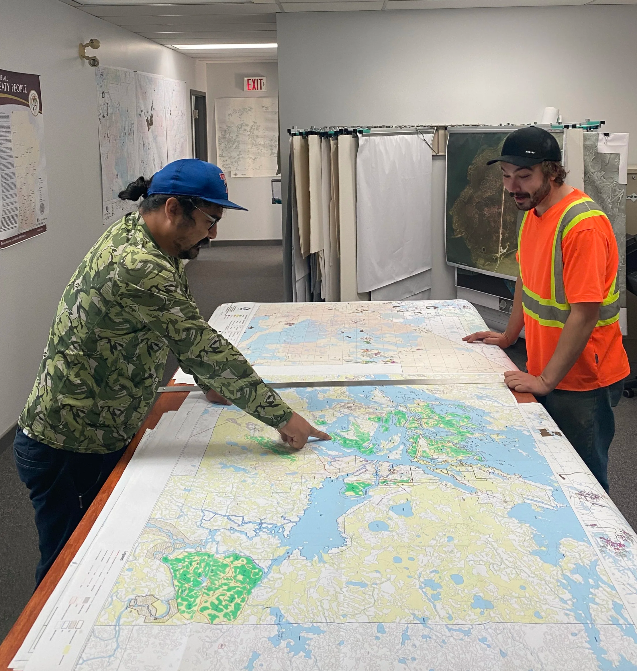 Two men review a large map spread out on a table in an office, with one pointing at the map and the other looking on, while discussing the map.