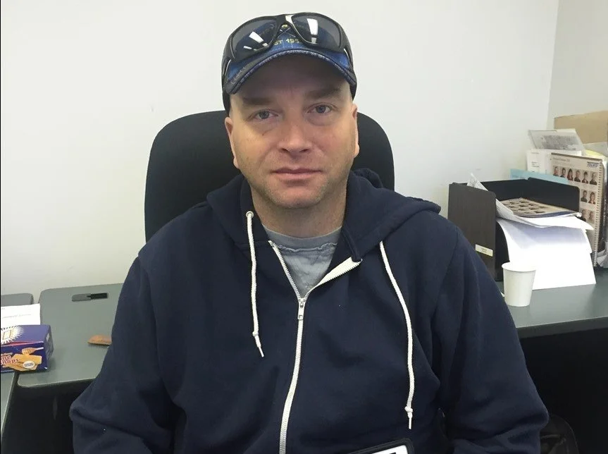 A man wearing a navy hoodie, gray t-shirt, and a baseball cap with sunglasses on top, sitting at a desk in an office.
