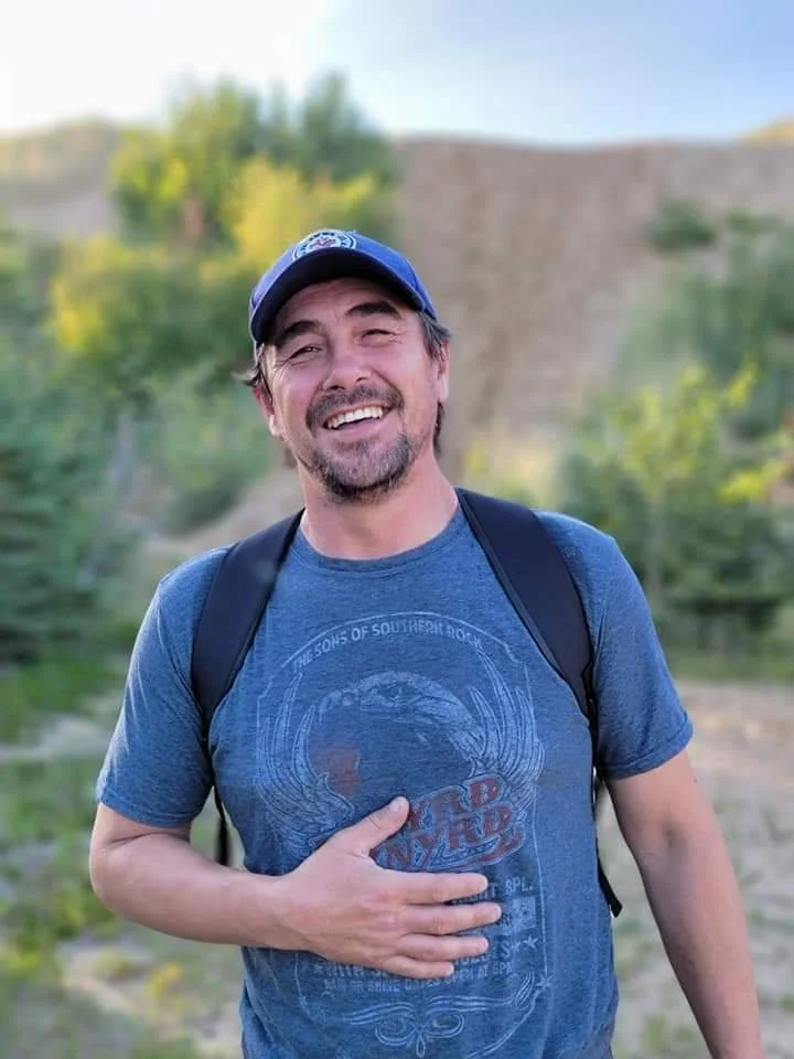 Man smiling outdoors wearing a blue T-shirt and a backpack, with a natural background of trees and a stone wall.