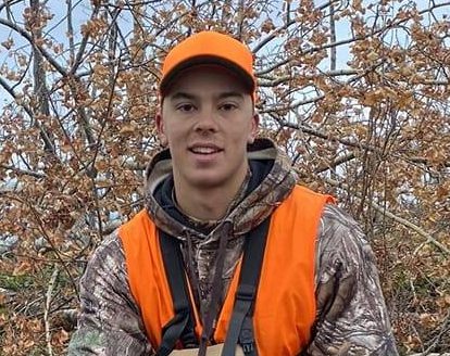 Young man in camouflage jacket and orange safety vest with orange cap, outdoors with trees and bare branches.