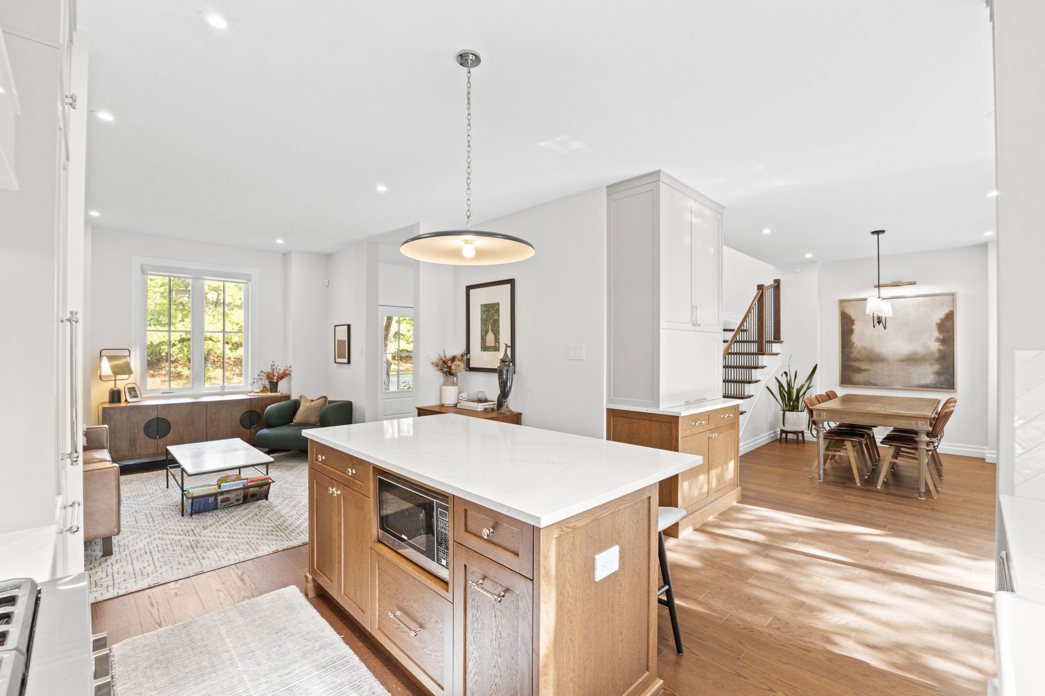 Open-plan living room and dining area in a modern home with white walls, hardwood floors, a kitchen island with a microwave, a sitting area with a green sofa, wooden furniture, and large windows letting in natural light.
