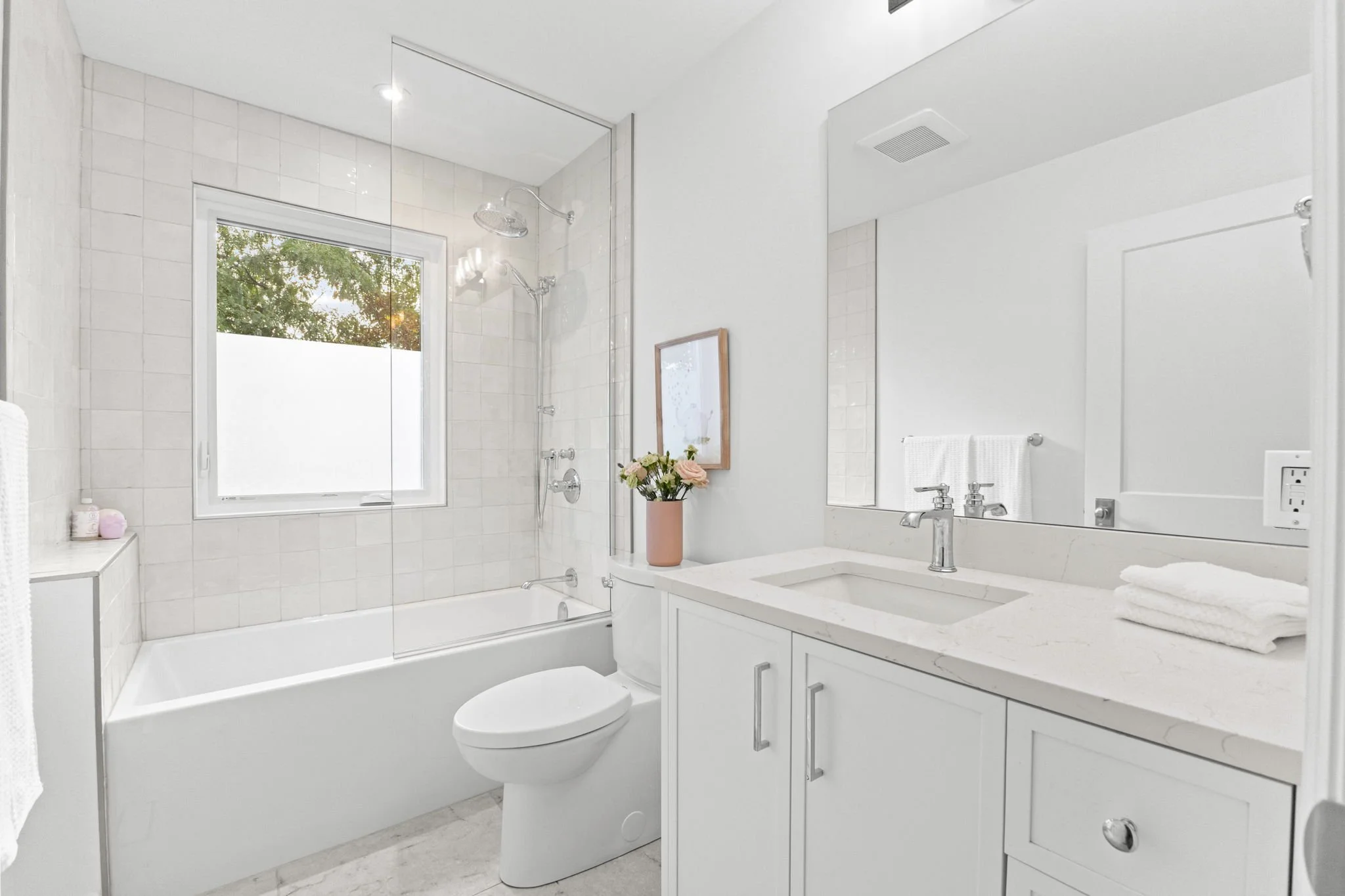 Bright, modern bathroom with a bathtub and shower, white tiles, a window, a white vanity with a sink, a large mirror, folded towels, and a decorative flower arrangement.