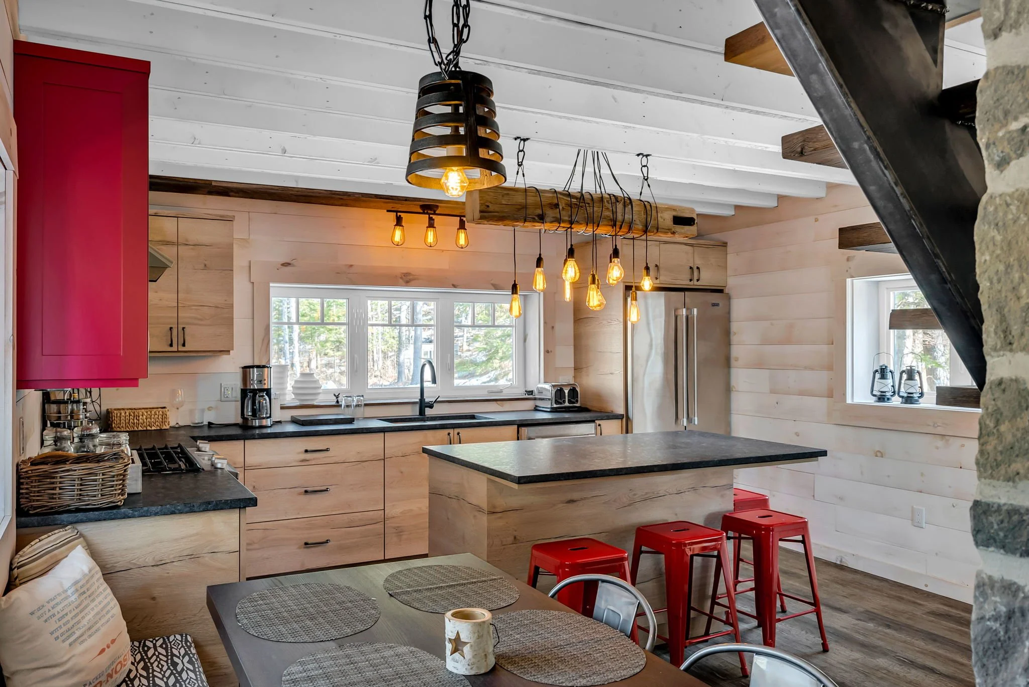 Big Rideau Lake cottage bunkie kitchen with light wood cabinets, black countertops, stainless steel appliances, island with seating, and Edison bulb pendant lights in a cozy cabin setting by Centennial Construction Management.