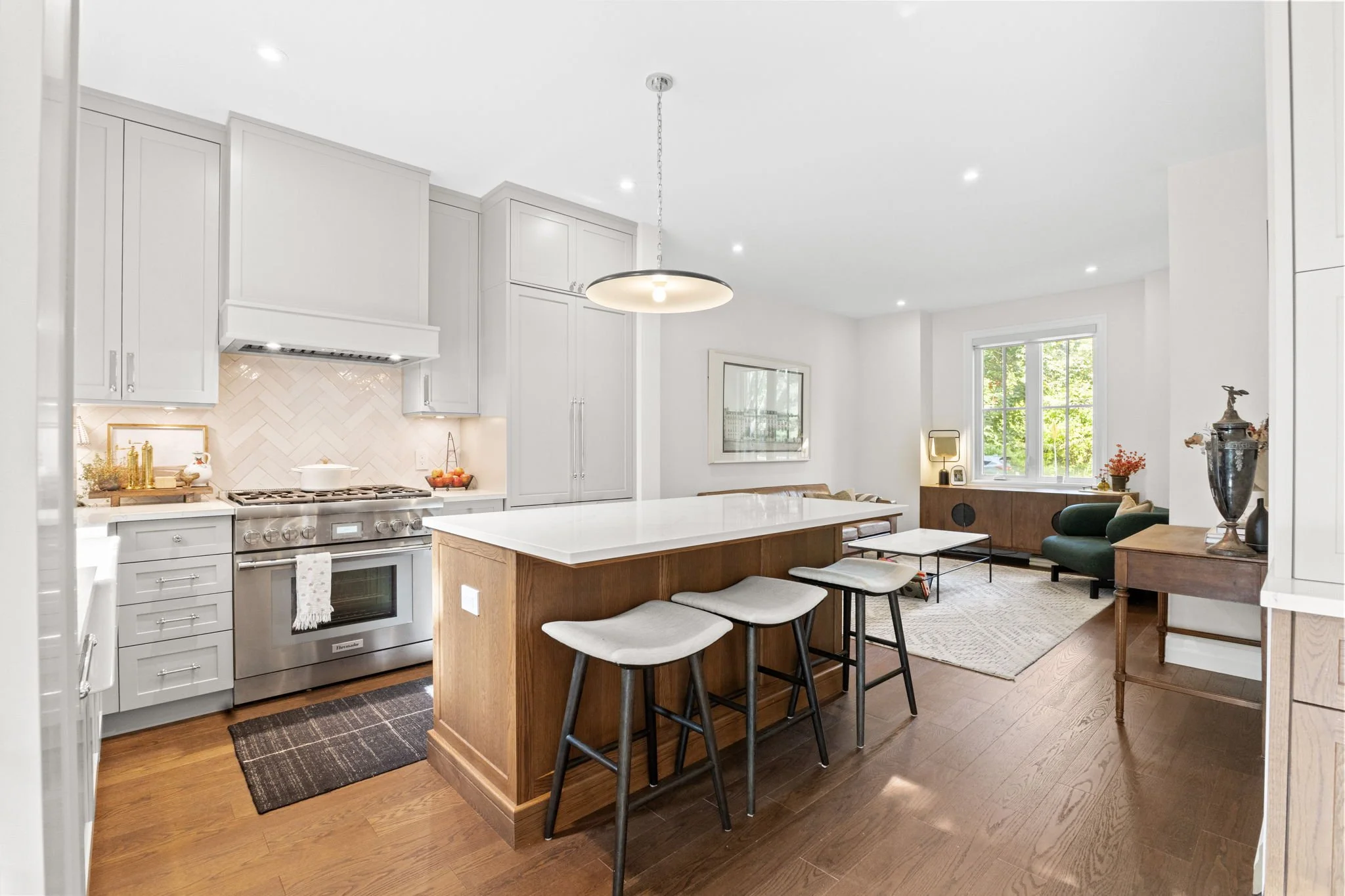 Open-concept kitchen and living room with white cabinets, stainless steel stove, island with white countertop and black barstools, hardwood floors, and a sitting area with green and wooden furniture near windows.