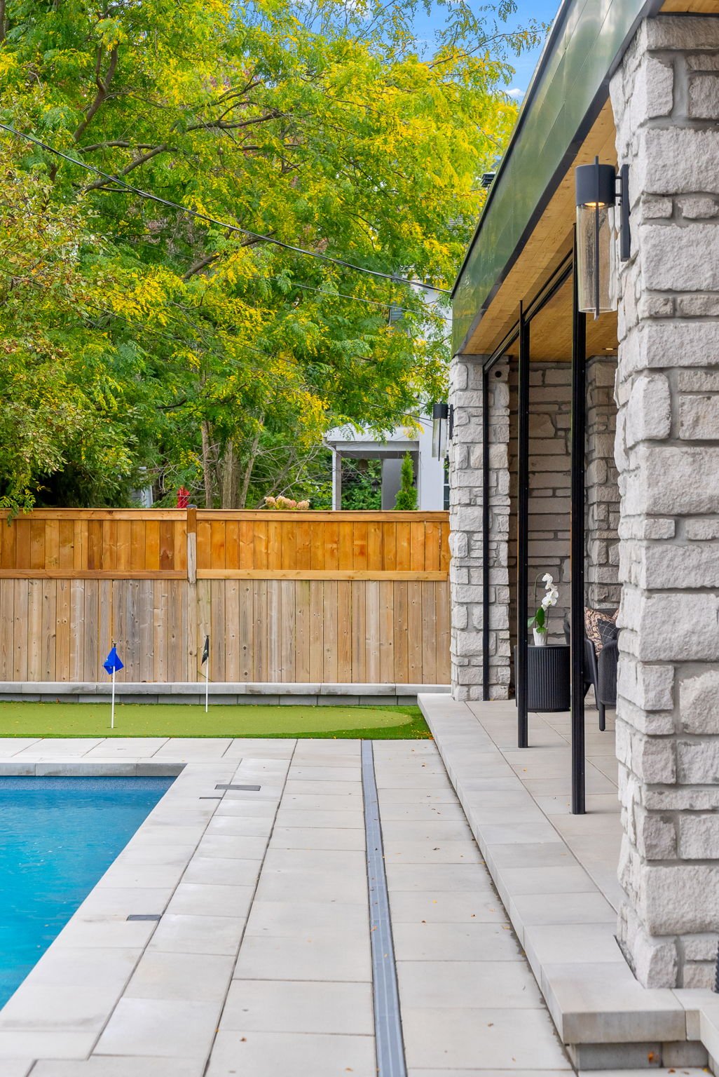 A backyard with a swimming pool, a wooden privacy fence, green trees, a tiled patio, and a portion of a house with stone walls and black framed glass doors.