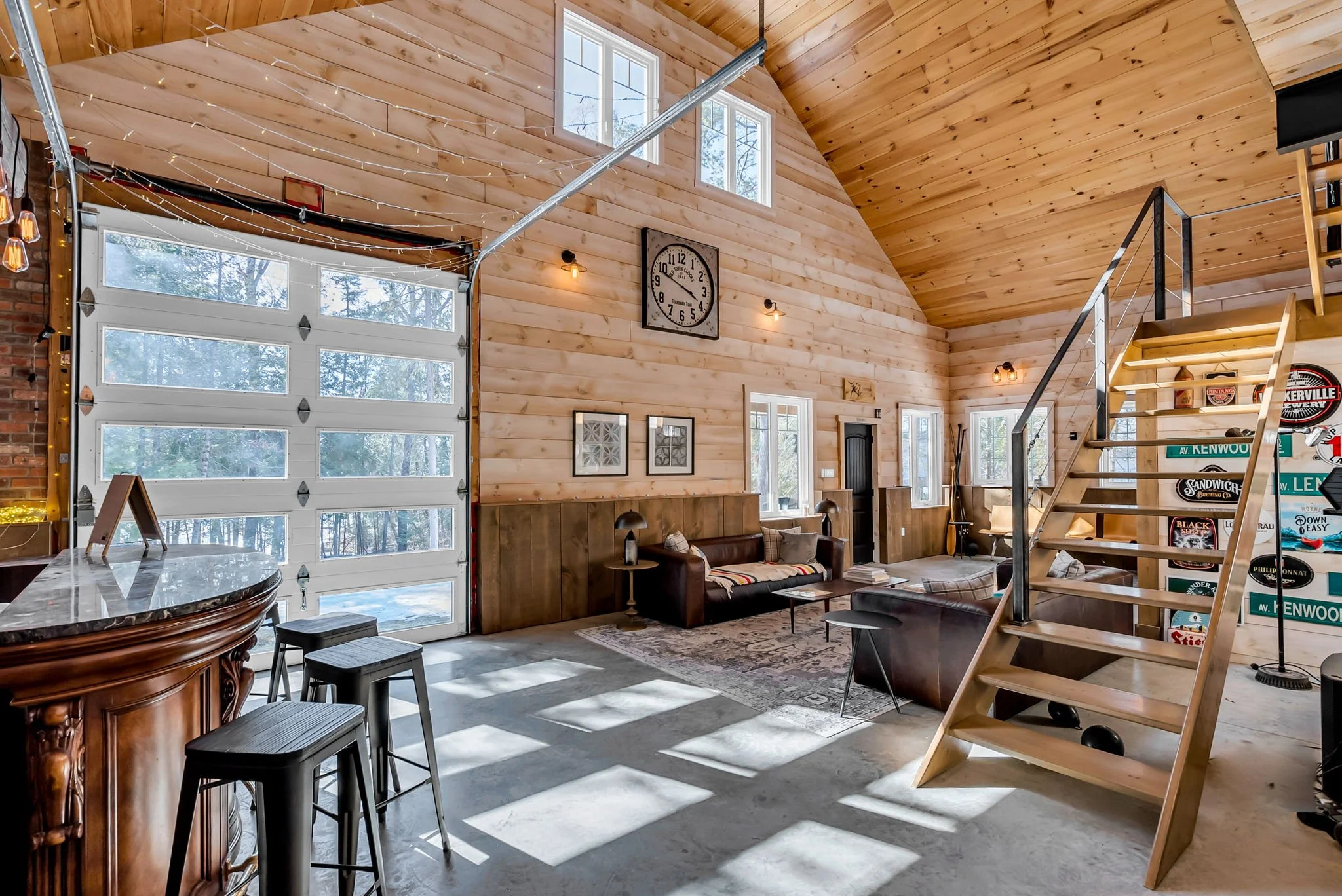 Interior of a rustic, modern living room with a high, vaulted wooden ceiling, wood-paneled walls, and a concrete floor. Features include a large garage-style door on the left, a black sofa with throw pillows, a patterned area rug, and a staircase lea