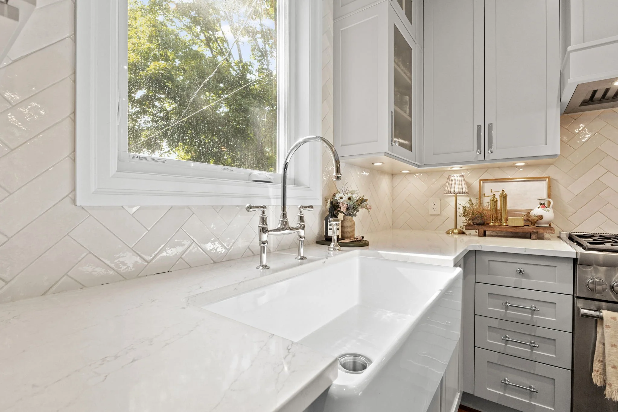 Modern kitchen sink with white marble countertop, gray cabinets, window with green tree outside, decorative items including flowers, lamp, and framed picture on the counter.