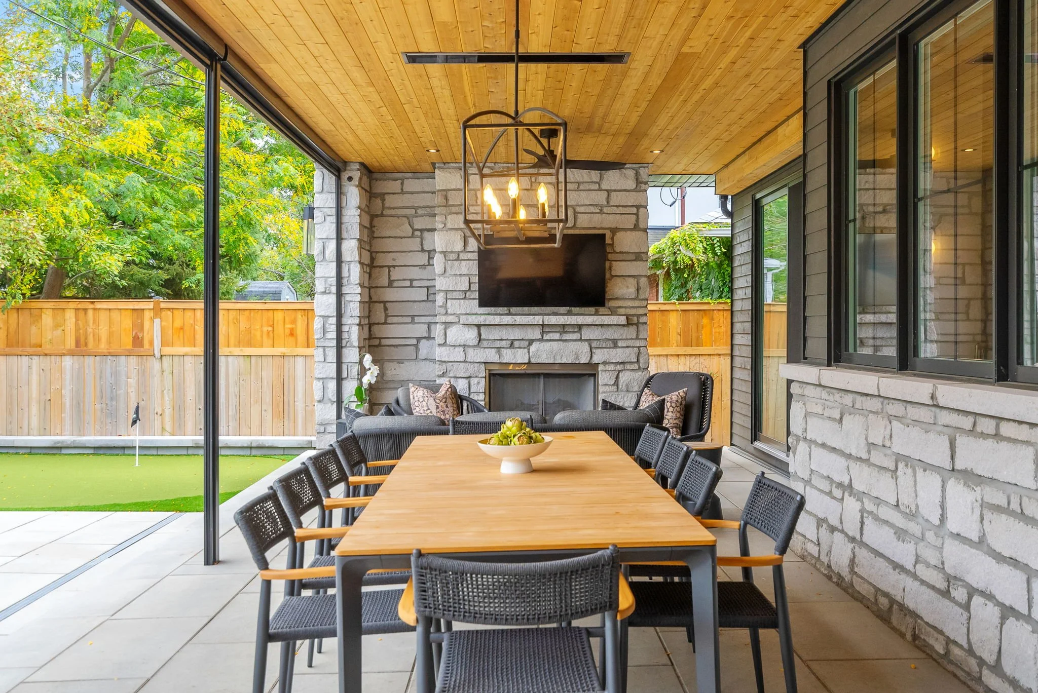 Covered outdoor patio with black dining table, black chairs with wooden arms, a stone fireplace, a wall-mounted TV, and a ceiling light fixture. Green yard with trees and a wooden fence in the background.