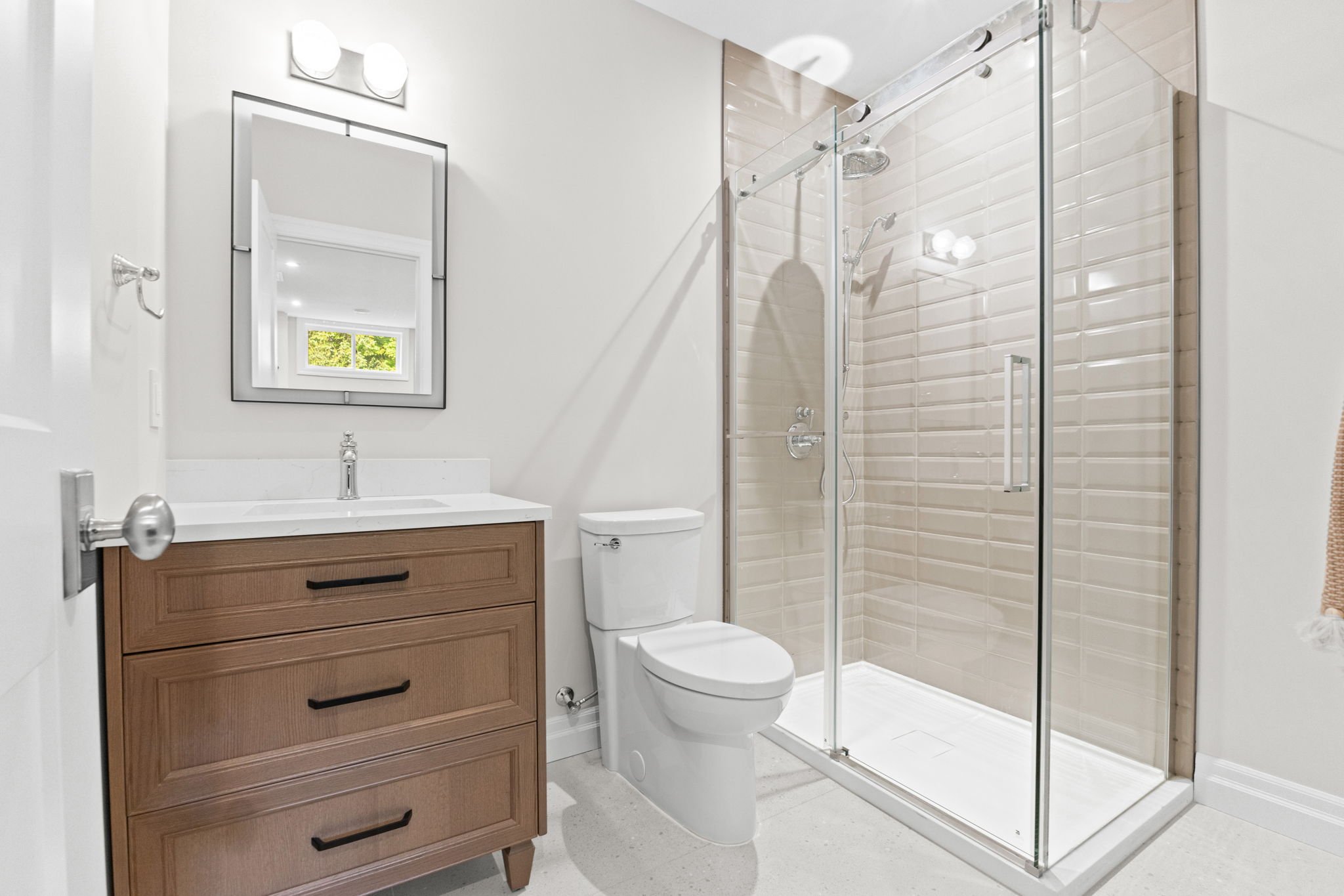 A modern bathroom with a wooden vanity, white countertop, mirror, toilet, and a glass-enclosed shower with beige tiles.