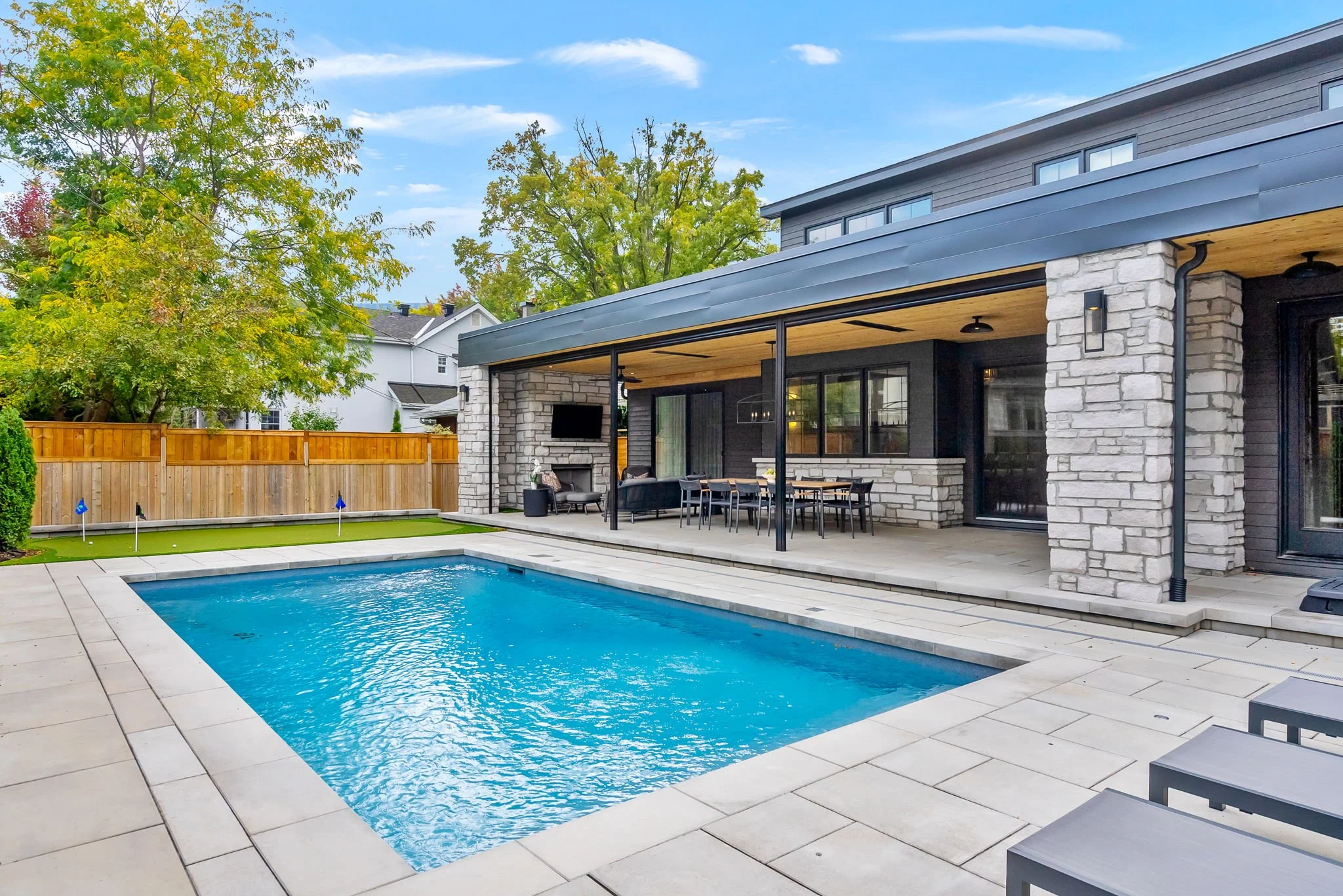 Backyard with a swimming pool, patio, outdoor furniture, and a stone fireplace, surrounded by a wooden fence and trees.