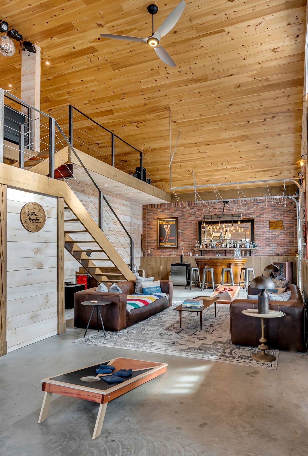 Interior of a modern lounge with a high wooden ceiling, brick walls, leather sofas, a bar area with stools, and a loft space above.