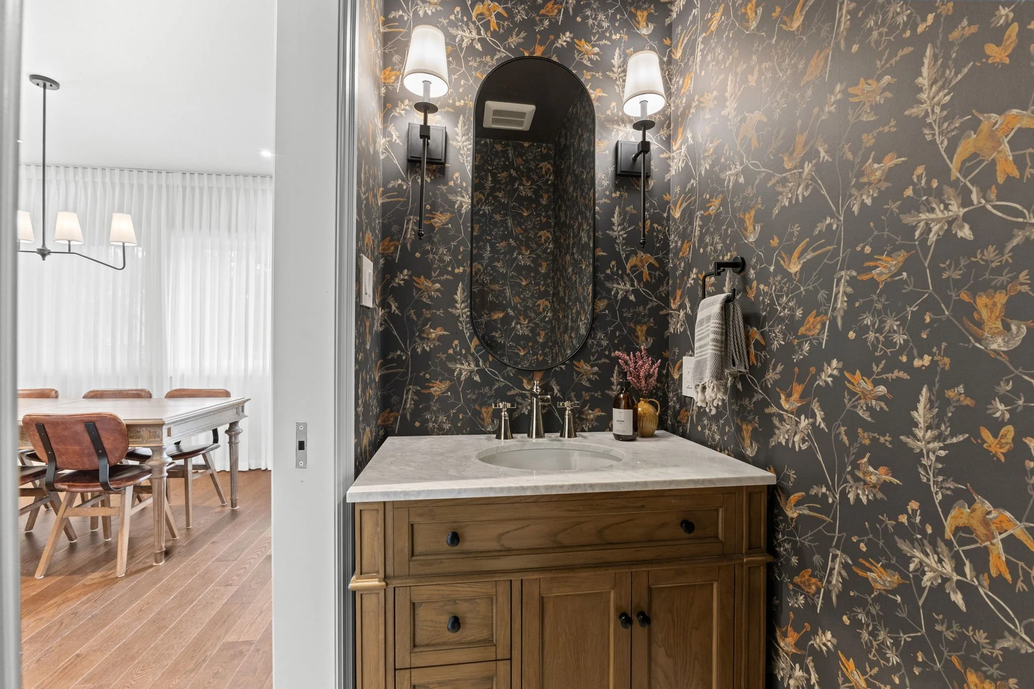 A bathroom vanity with a marble countertop, a mirror with an oval shape, wall sconces, a towel hook with a gray towel, and decorative items including a bottle, a vase with pink flowers, and a soap dispenser.