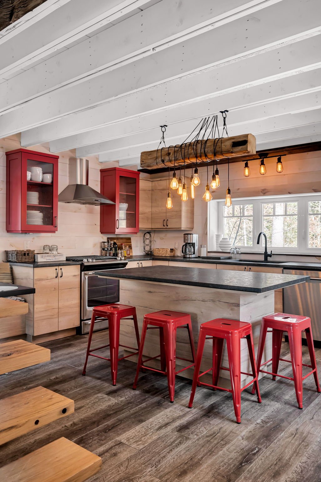 Rustic kitchen with wooden cabinets, black countertops, red bar stools, and an island. Features a modern stove, hanging Edison bulbs, and a large window overlooking trees.