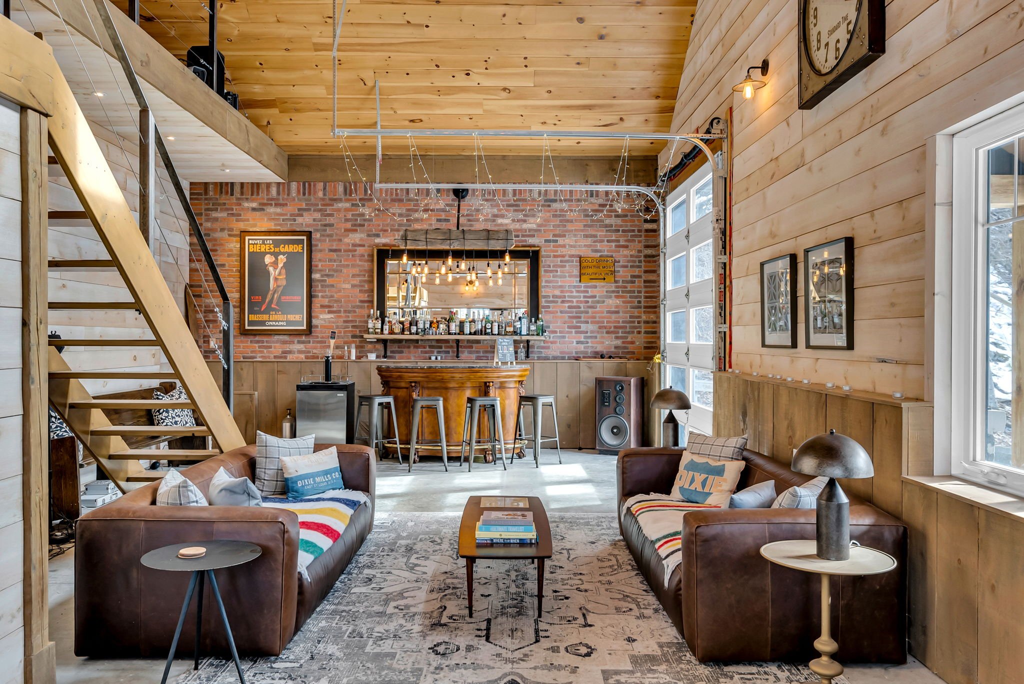 A cozy living room with wooden walls and a staircase on the left. There are two brown leather sofas with colorful blankets and pillows, a small side table with a lamp on the right, and a rectangular coffee table in the center. The back wall features 