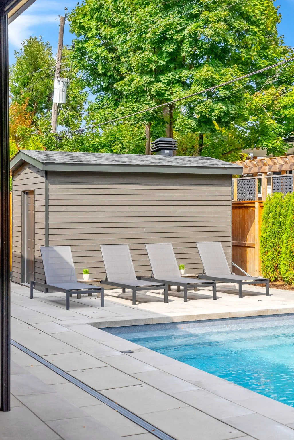 Swimming pool with four lounge chairs and small potted plants on a patio, surrounded by a wooden fence, trees, and a gray shed.