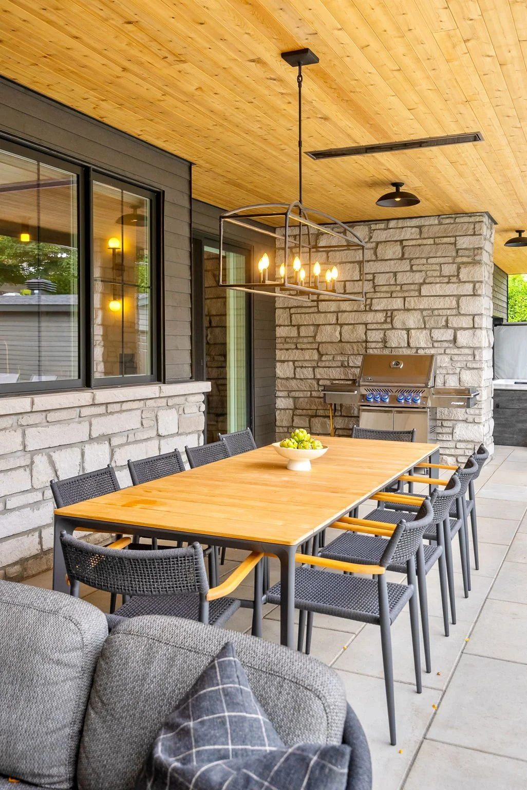 Outdoor patio with a wooden dining table, ten black chairs, a bowl of green grapes, a metal chandelier with multiple lights, a propane grill, stone and wood walls, large windows, and a wooden ceiling.