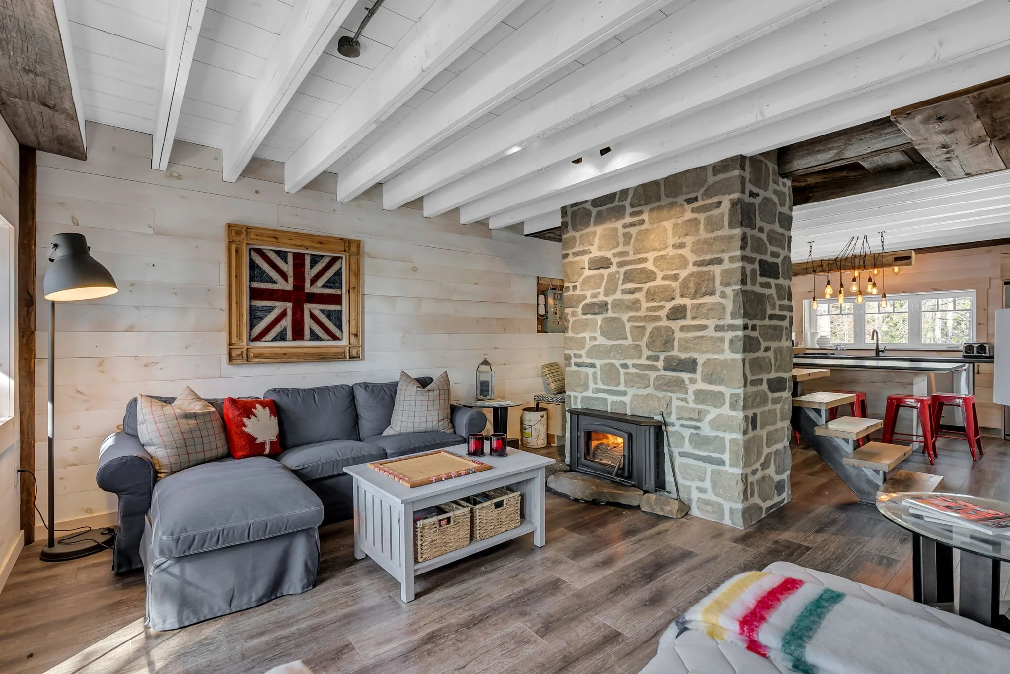 Living room with a gray sofa, wood stove, white wooden walls, stone chimney, and kitchen in the background with red stools.