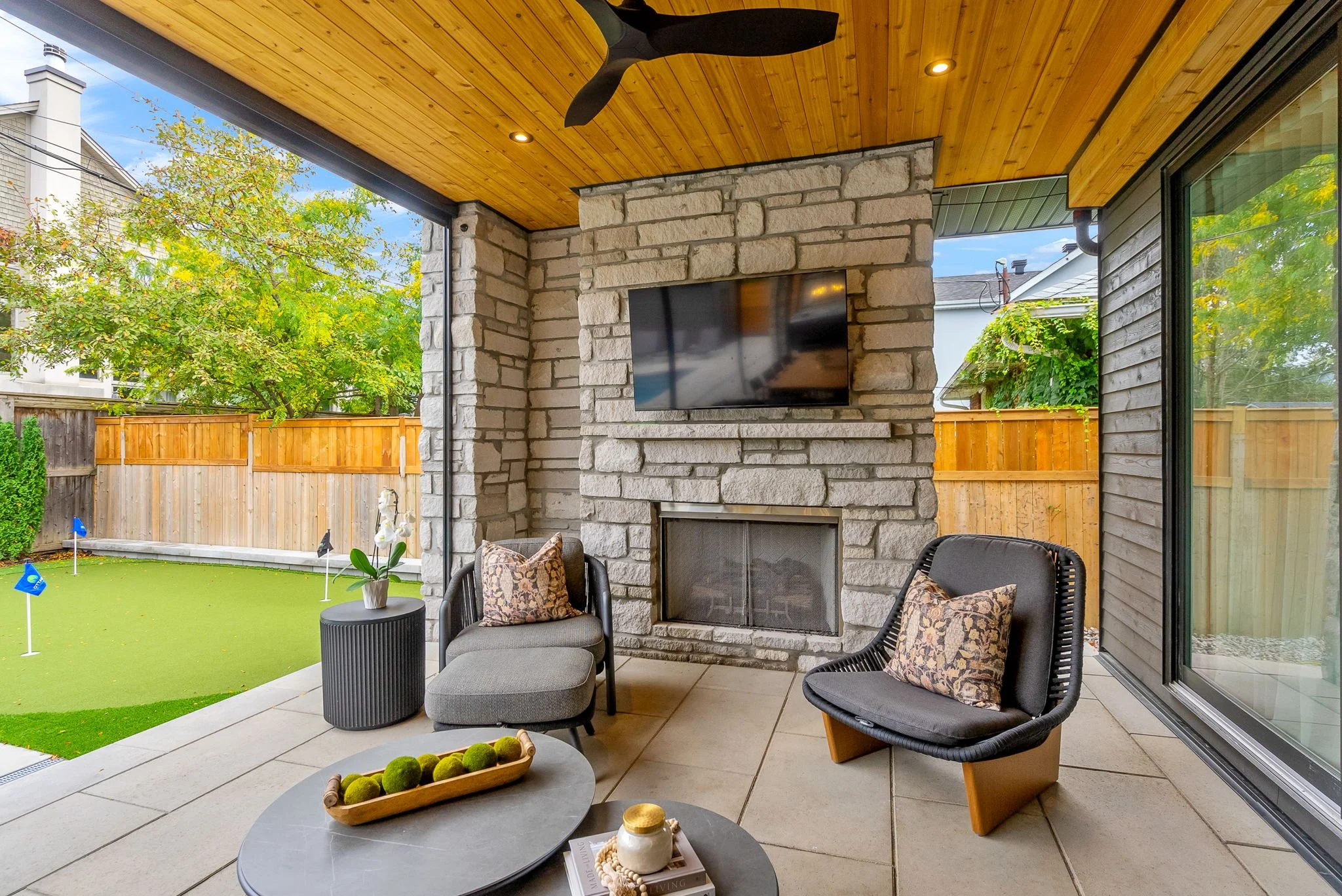 Covered patio area with two modern black chairs with cushions, a gray ottoman, and a gray side table with a decorative bonsai tree, overlooking a backyard with grass golf holes and a wooden fence, and a stone fireplace with a mounted TV above and a c